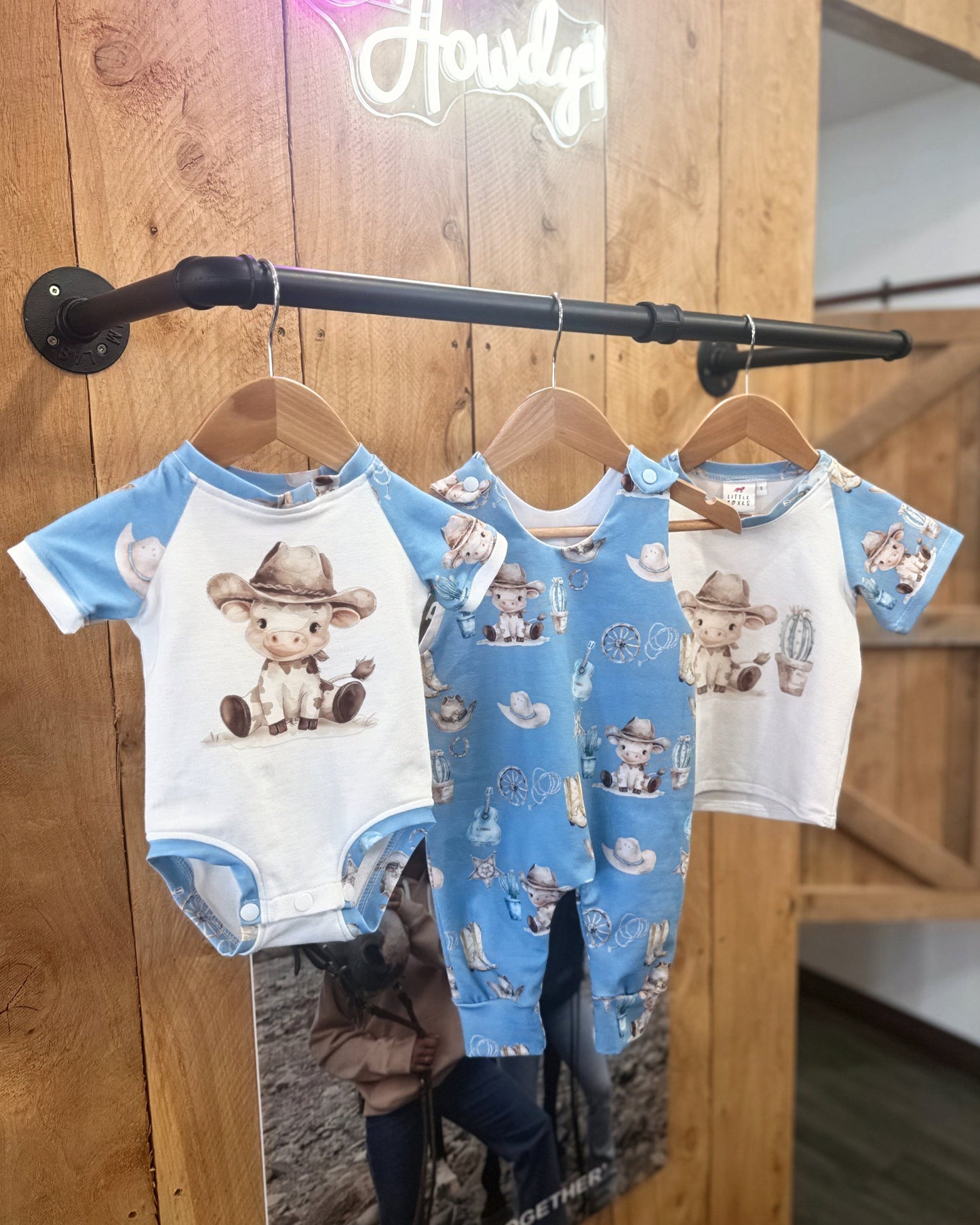 Three baby rompers with cowboy design hanging on a rack against a wooden wall.