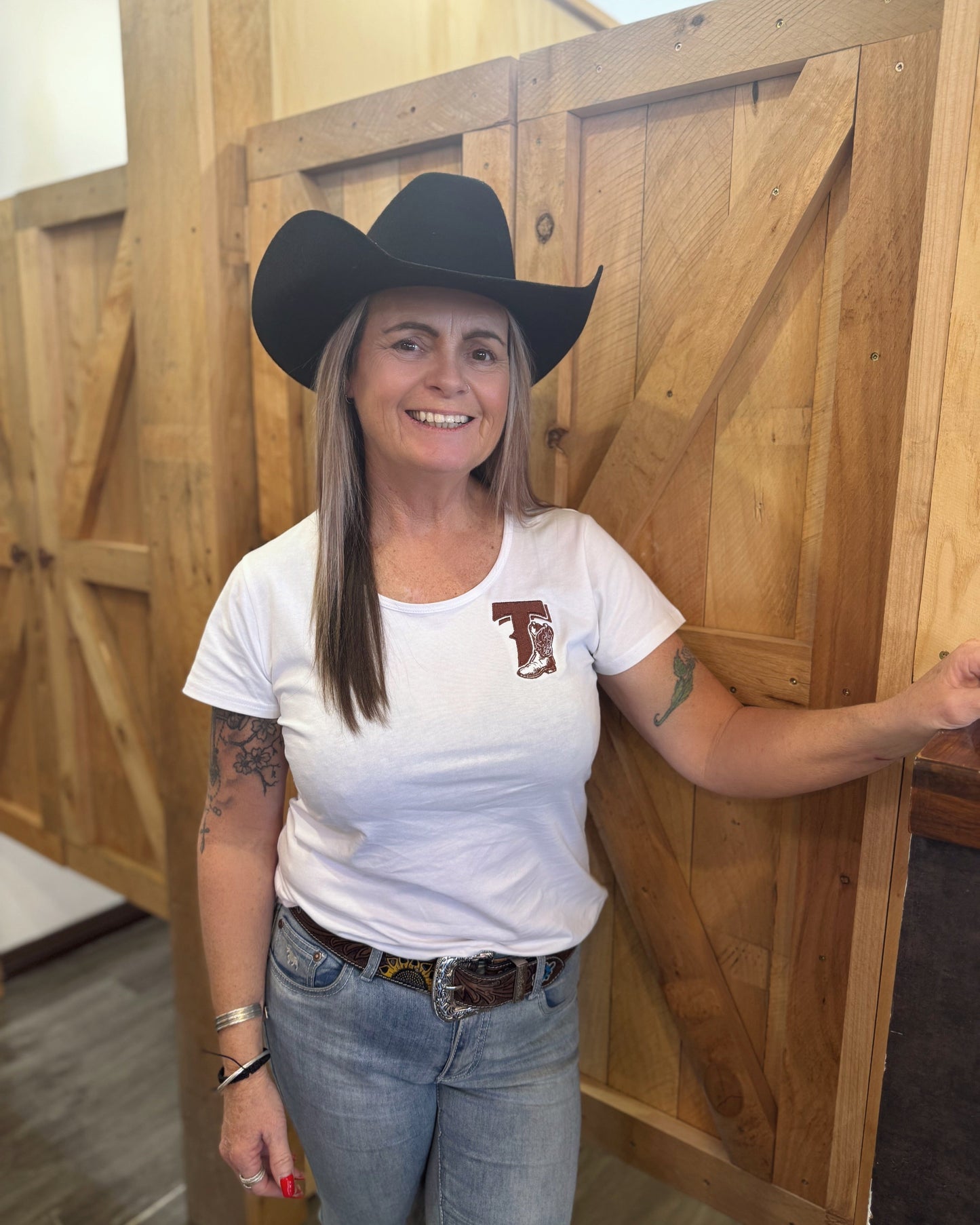 Woman wearing a cowboy hat and white t-shirt with a logo, standing in front of wooden doors.