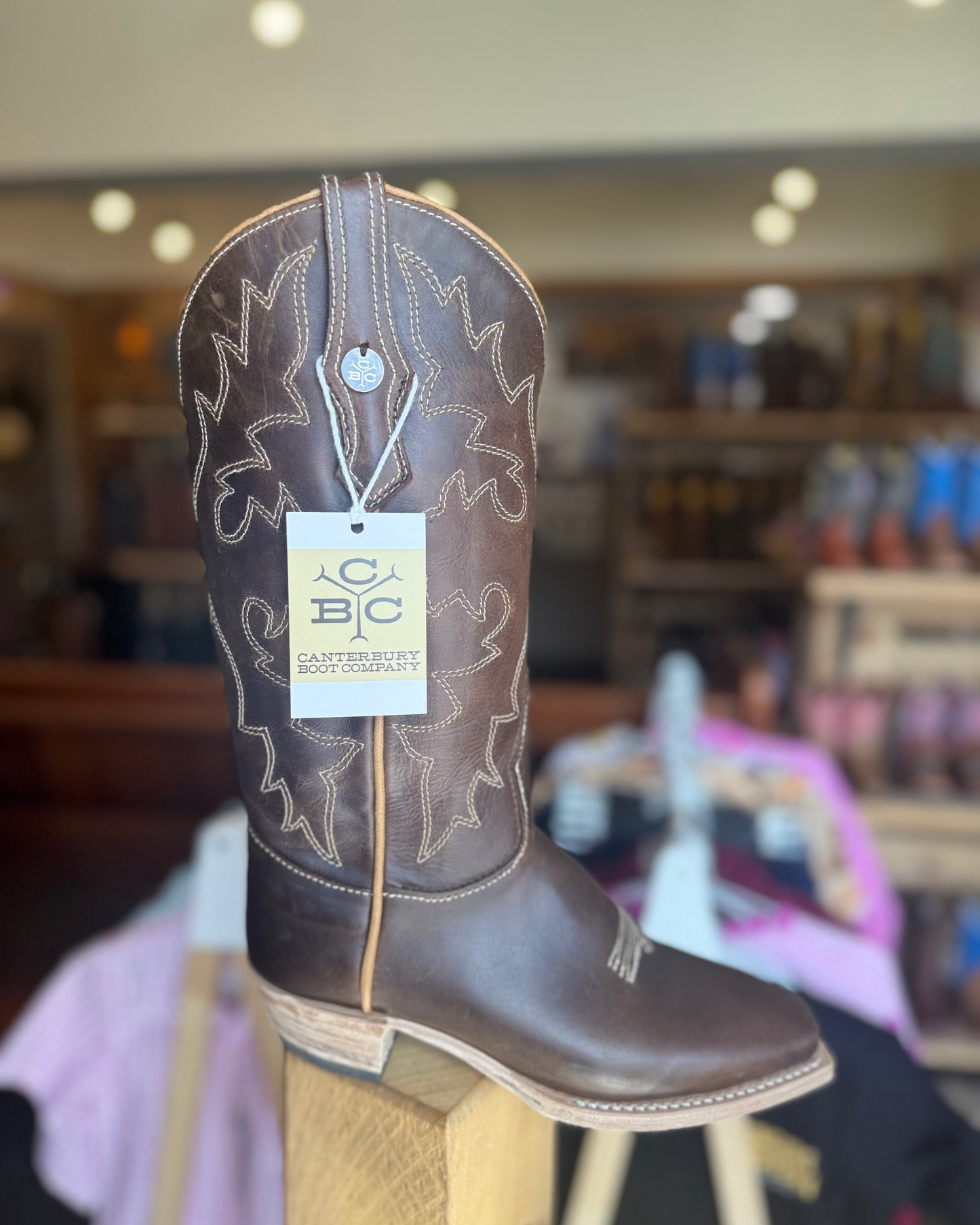 Brown cowboy boot on a wooden block with a visible brand tag in a store setting.
