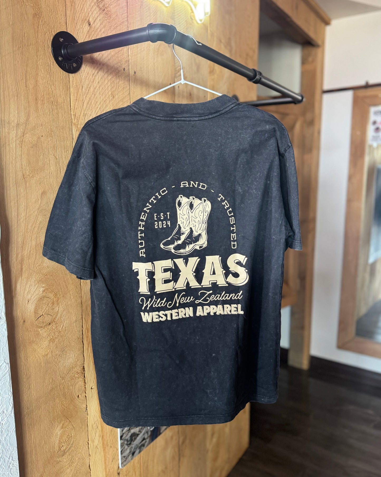Black t-shirt with 'Texas' and boot design on a hanger against a wooden wall.