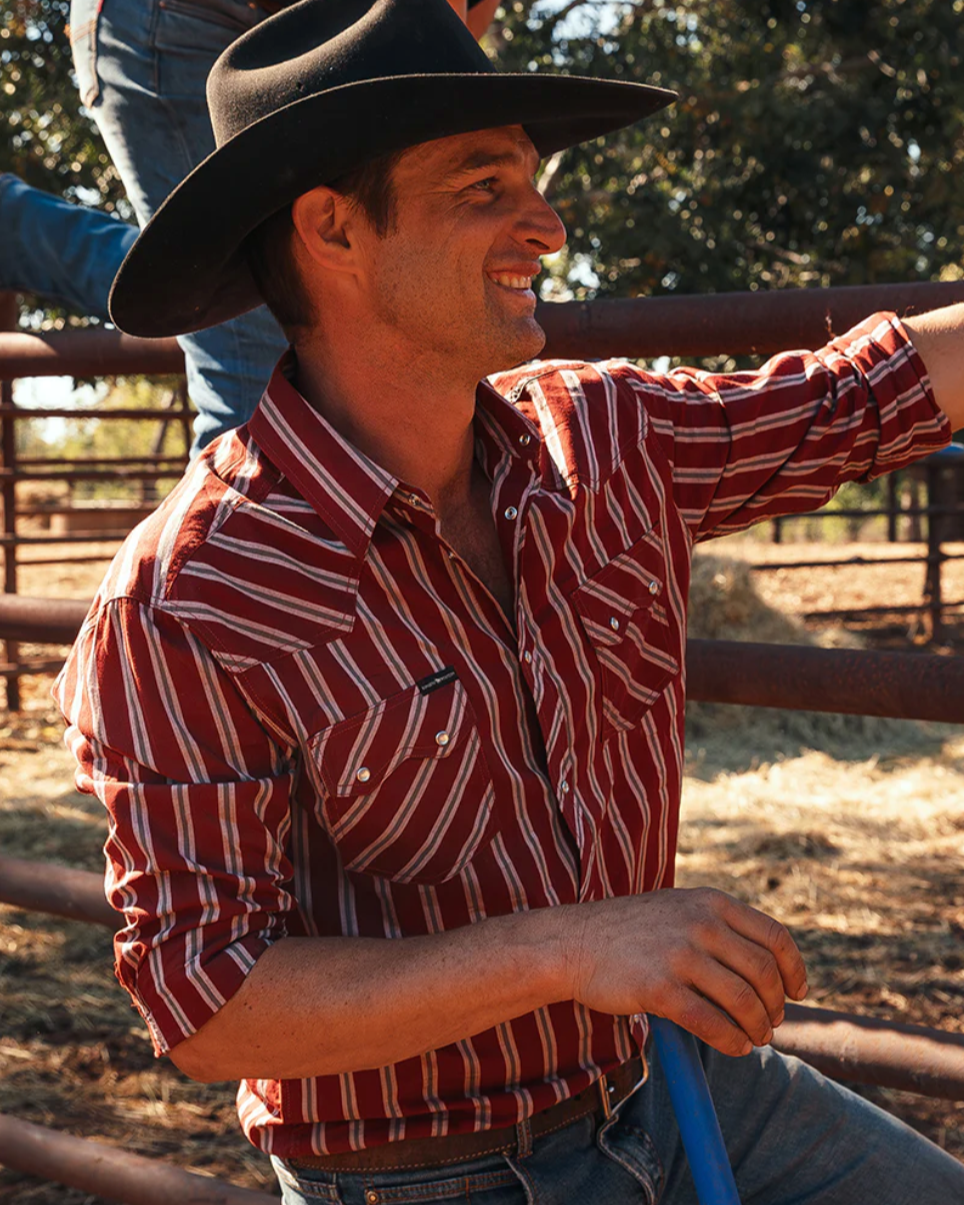 Man wearing a cowboy hat and red striped shirt standing in a rural setting.