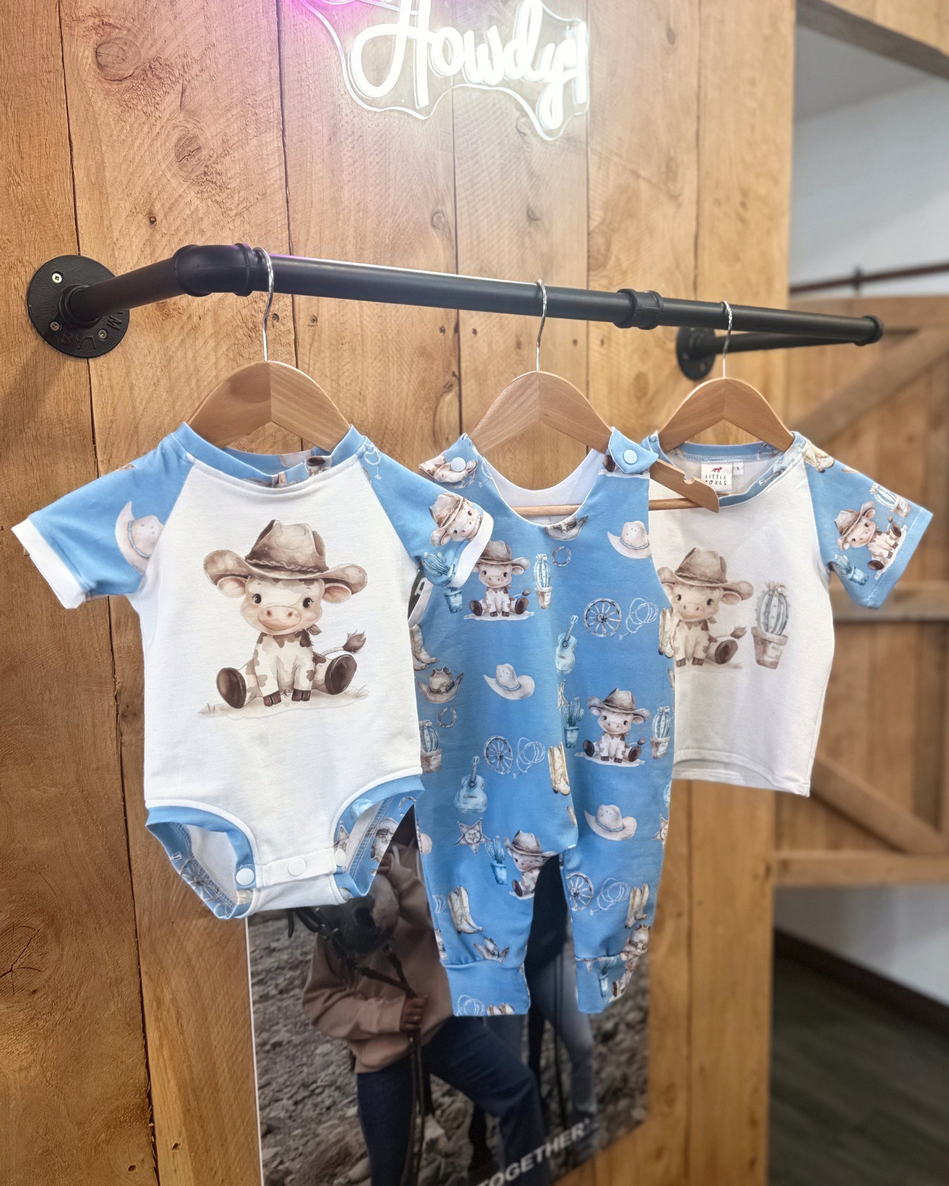 Three baby rompers with cowboy design hanging on a rack against a wooden wall.