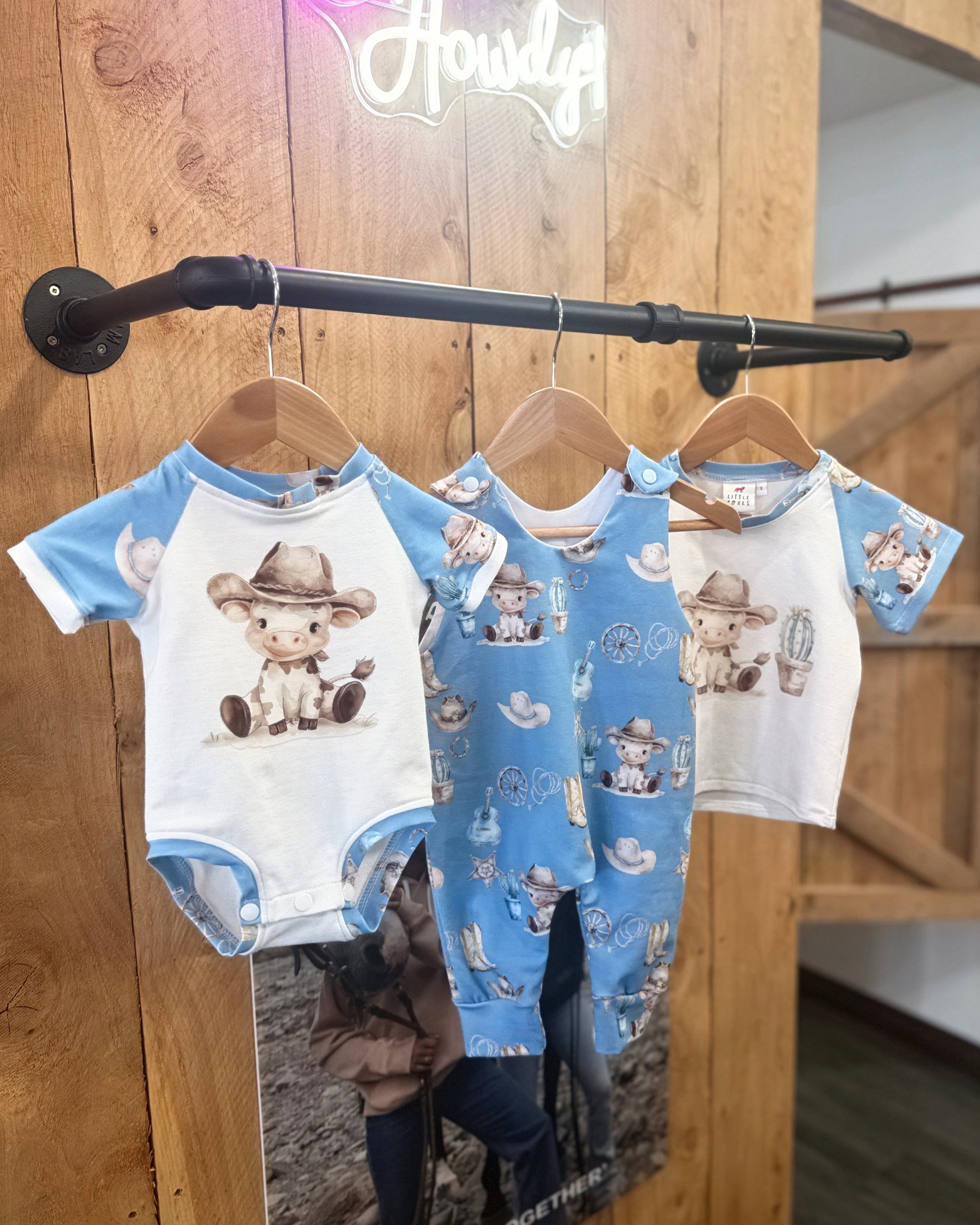 Three baby rompers with cowboy design hanging on a rack against a wooden wall.