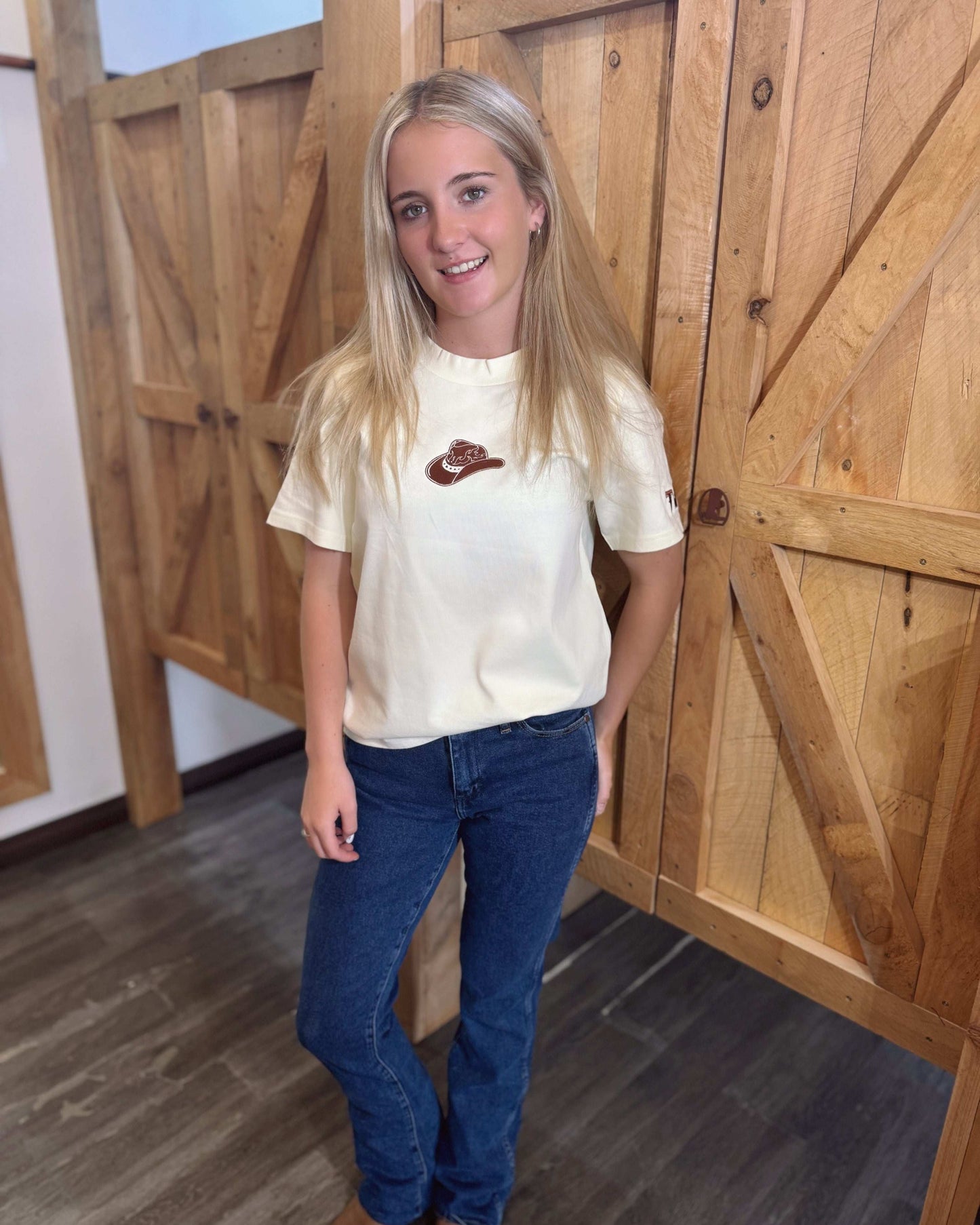 A butter yellow women's Texas Western Apparel T-shirt with a classic western style. The front features an embroidered cowboy hat, while the sleeve is detailed with "TWA" initials, adding a subtle branded touch.