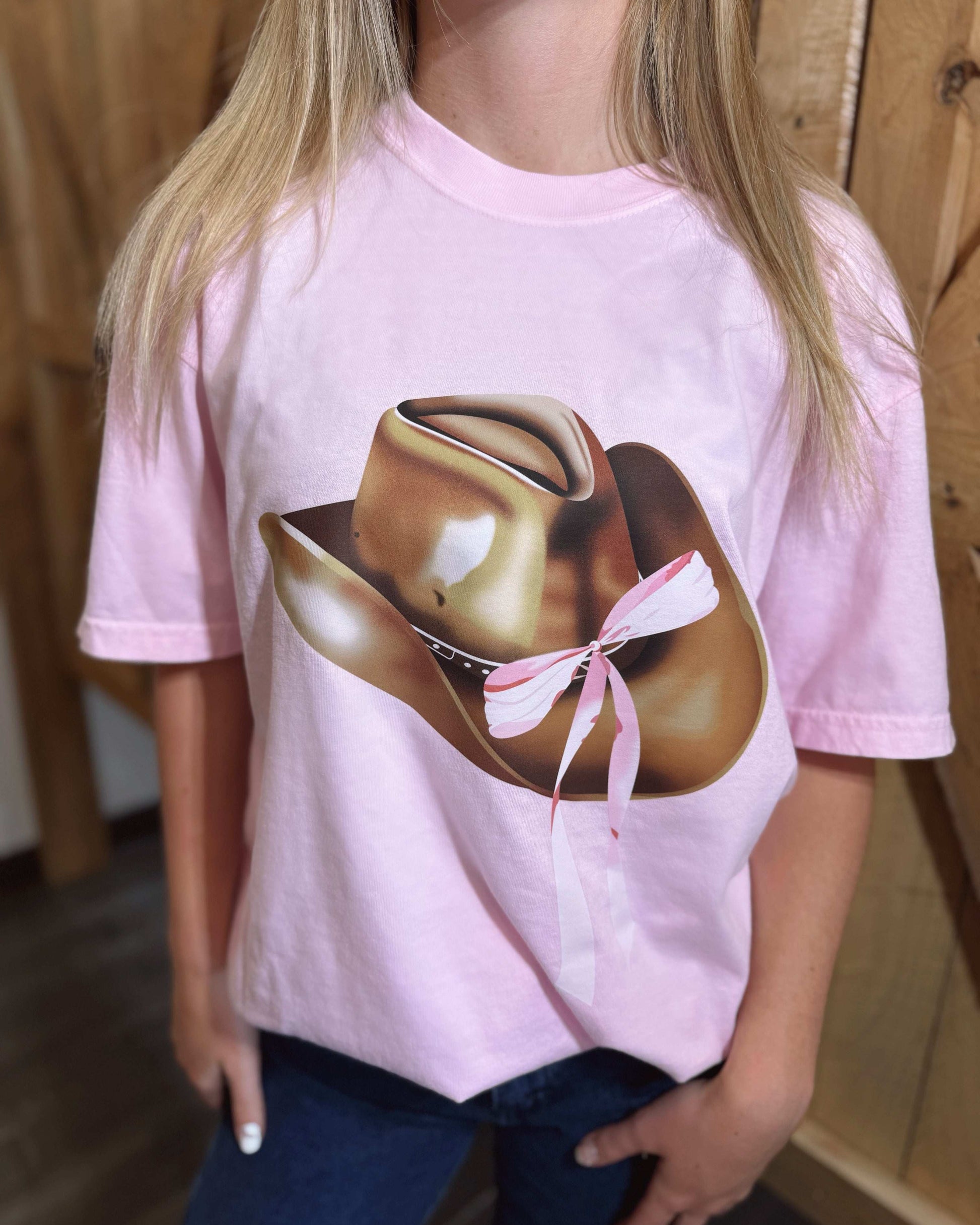 A pastel pink oversized T-shirt featuring a bold graphic print of a metallic gold cowboy hat with a pink and white ribbon tied around it. The shirt has a relaxed fit, short sleeves, and a classic crew neckline. The back of the shirt is plain pink. Modeled with dark blue jeans against a rustic wooden background.