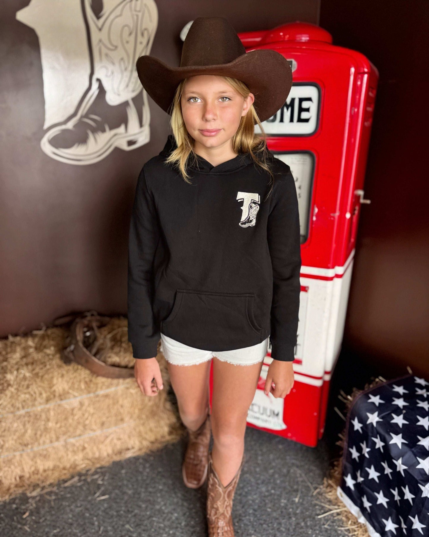 Black Texas Western Apparel youth/kids hoodie with detailed embroidery. The front features a stitched 'T' and a Western-style boot, while the back displays the Texas Western Apparel logo. Perfect for little cowboys and cowgirls who love bold Western style!