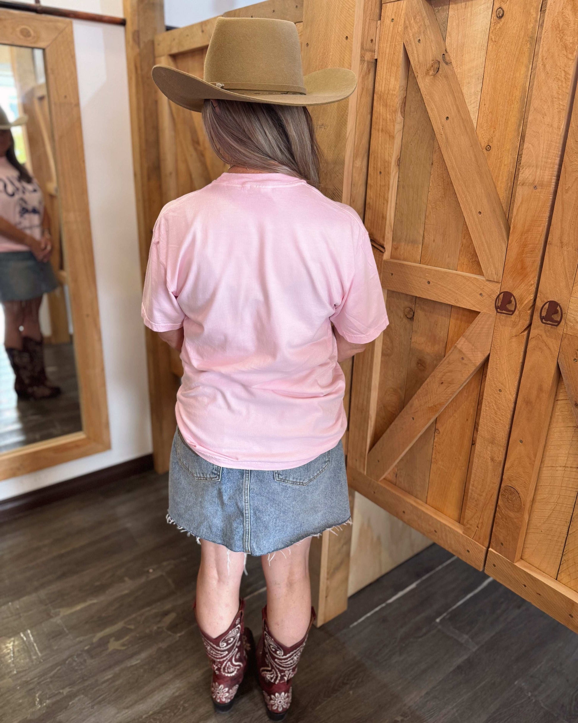 A woman with long, wavy hair wears a pastel pink oversized t-shirt with bold navy blue text that reads "TEXAS" along with a galloping horse graphic and the words "EST" and "Now That's Country." She pairs the shirt with a frayed denim mini skirt, a wide-brimmed beige cowboy hat, and embroidered cowboy boots. From the back, showing the plain pink back of the shirt with a relaxed fit.