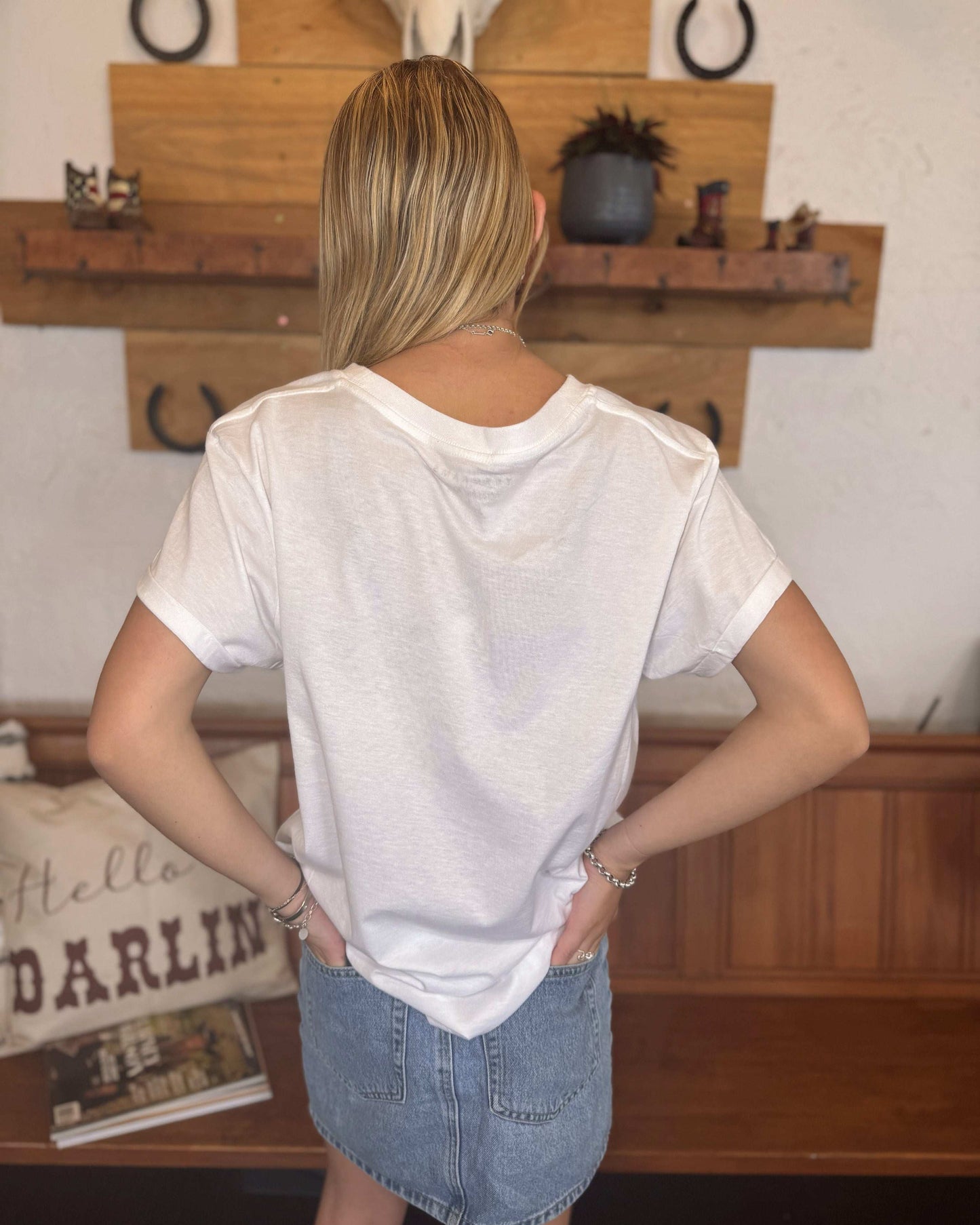 A young woman with long blonde hair wears a white t-shirt featuring the phrase "Dirty Martini Please" in bold, blocky, black letters. She pairs it with a light blue denim mini skirt. The setting includes a wooden bench, a "Hello Darling" decorative pillow, and rustic wooden wall decor with horseshoes and a cow skull.