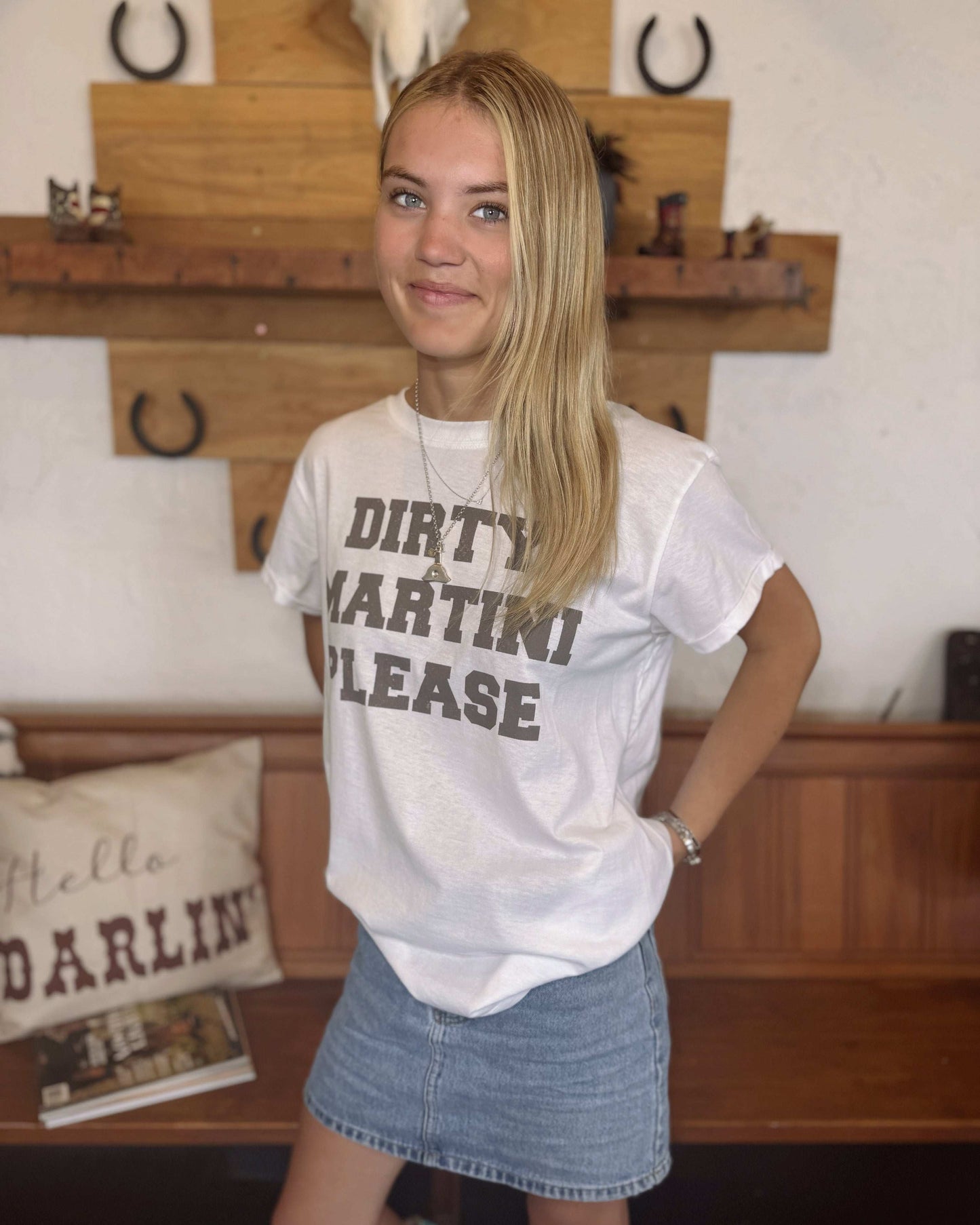 A young woman with long blonde hair wears a white t-shirt featuring the phrase "Dirty Martini Please" in bold, blocky, black letters. She pairs it with a light blue denim mini skirt. The setting includes a wooden bench, a "Hello Darling" decorative pillow, and rustic wooden wall decor with horseshoes and a cow skull.