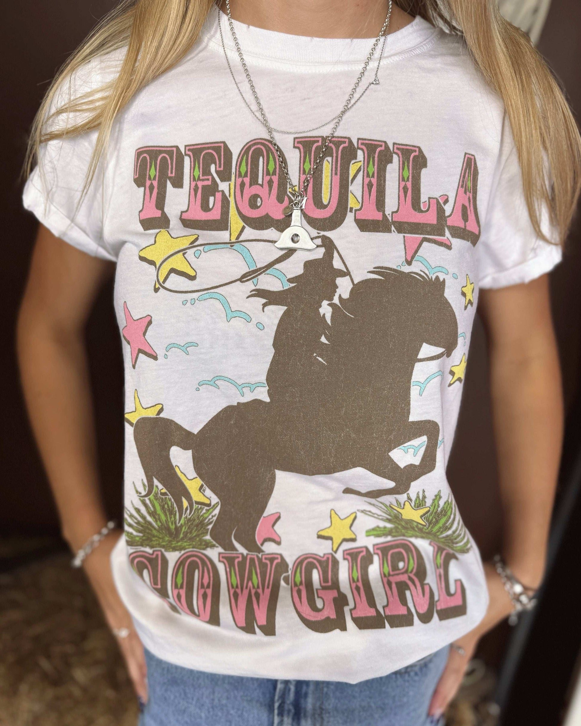 A close-up of a woman wearing a white graphic t-shirt featuring the words "TEQUILA COWGIRL" in bold, western-style pink and brown lettering. The design includes a silhouette of a cowgirl riding a rearing horse, surrounded by colorful stars, cacti, and swirling details. She is accessorized with layered silver necklaces, bracelets, and a silver ring. The t-shirt is slightly tucked into light-wash denim jeans. The background is blurred with a brown wall.