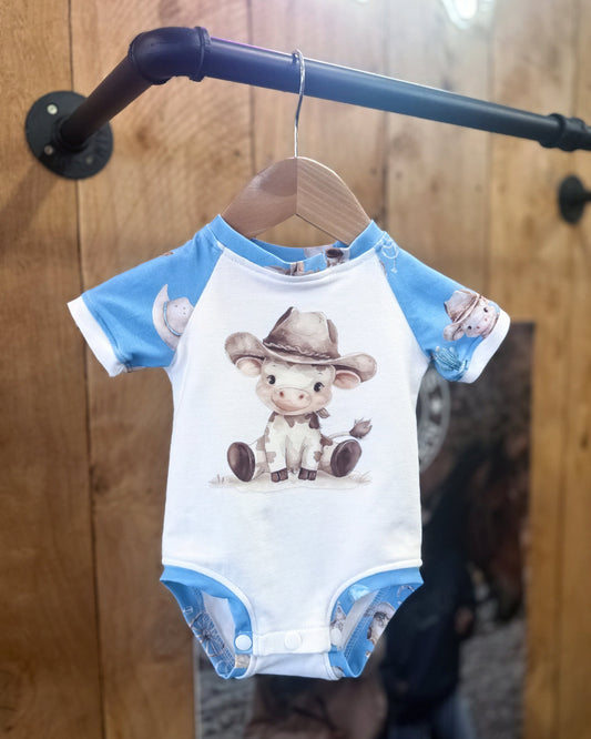 Baby onesie with cartoon cow design on a hanger against a wooden background