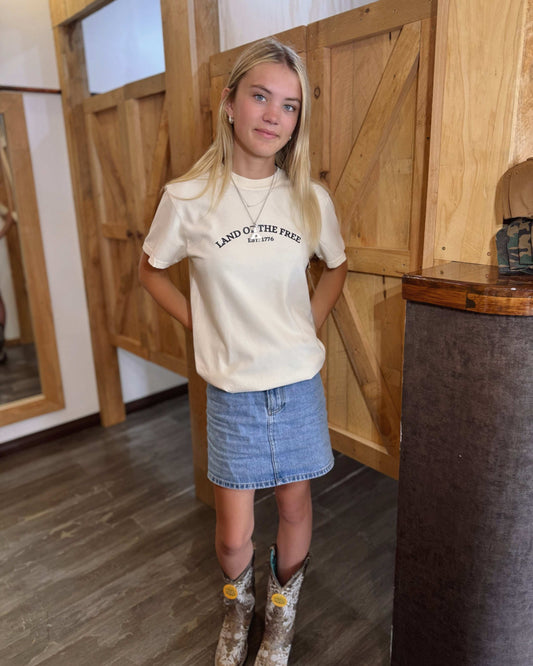 A young woman wears a cream-colored t-shirt with "Land of the Free Est. 1776" printed in black across the chest. The back showcases blue graphic illustrations of American icons, including the Empire State Building, pizza, and a food cart. She pairs it with a denim skirt and cowboy boots.