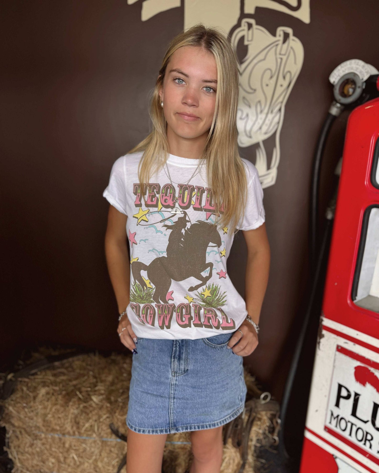 A close-up of a woman wearing a white graphic t-shirt featuring the words "TEQUILA COWGIRL" in bold, western-style pink and brown lettering. The design includes a silhouette of a cowgirl riding a rearing horse, surrounded by colorful stars, cacti, and swirling details. She is accessorized with layered silver necklaces, bracelets, and a silver ring. The t-shirt is slightly tucked into light-wash denim jeans. The background is blurred with a brown wall.