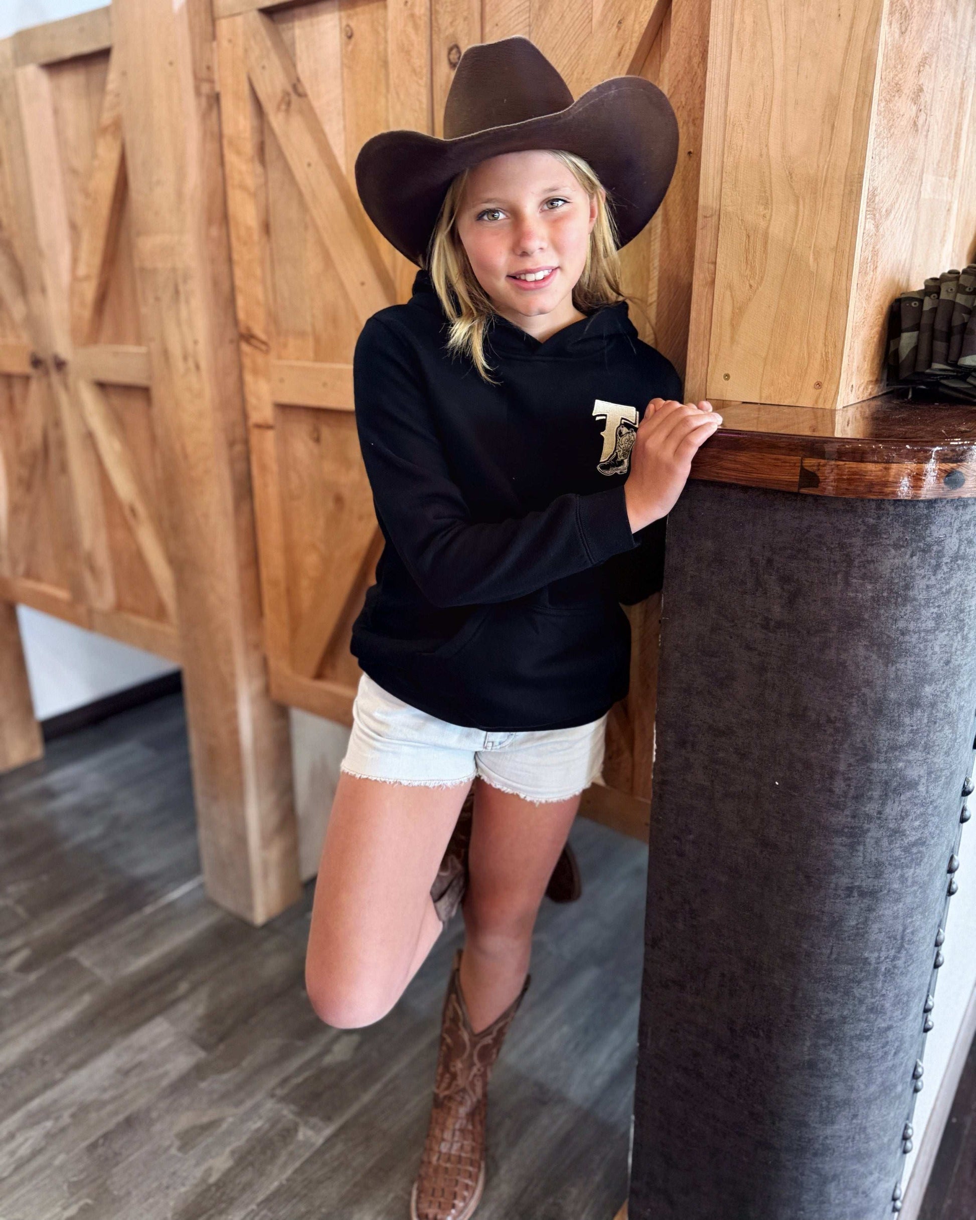 Black Texas Western Apparel youth/kids hoodie with detailed embroidery. The front features a stitched 'T' and a Western-style boot, while the back displays the Texas Western Apparel logo. Perfect for little cowboys and cowgirls who love bold Western style!