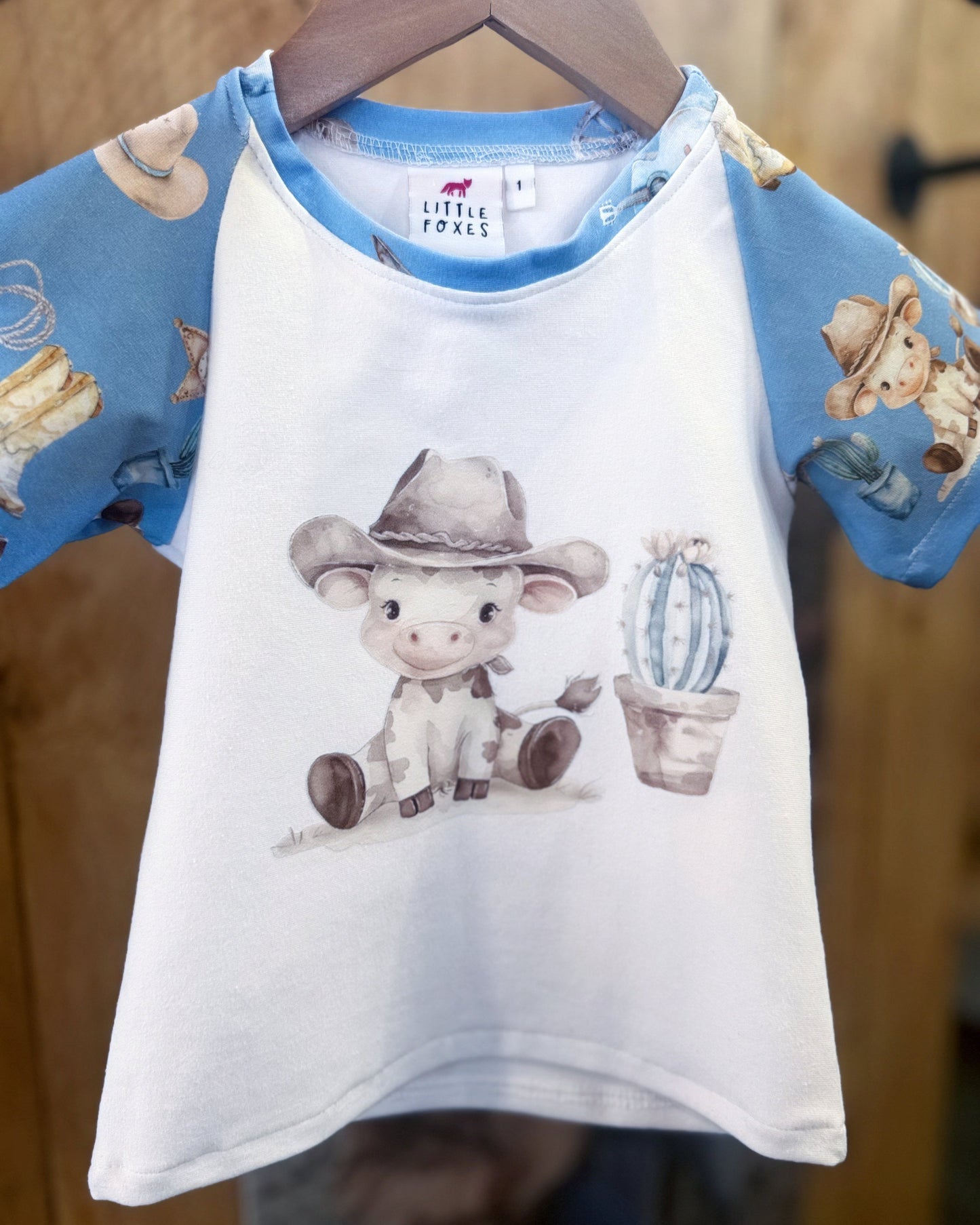 Children's shirt with cow print and blue sleeves on a hanger