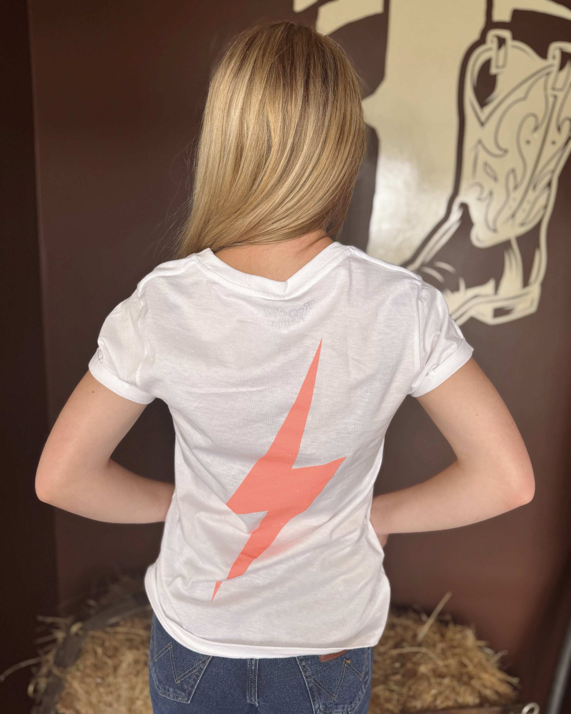 A woman wears a white graphic tee featuring bold red-orange text that reads, "AND SUDDENLY, ALL THESE SONGS WERE ABOUT YOU," with two stars at the bottom. The shirt has a relaxed fit and short sleeves, paired with classic blue jeans. The back of the tee showcases a striking red-orange lightning bolt design. The setting includes a rustic western-inspired backdrop with hay bales and vintage decor, complementing the casual country style.