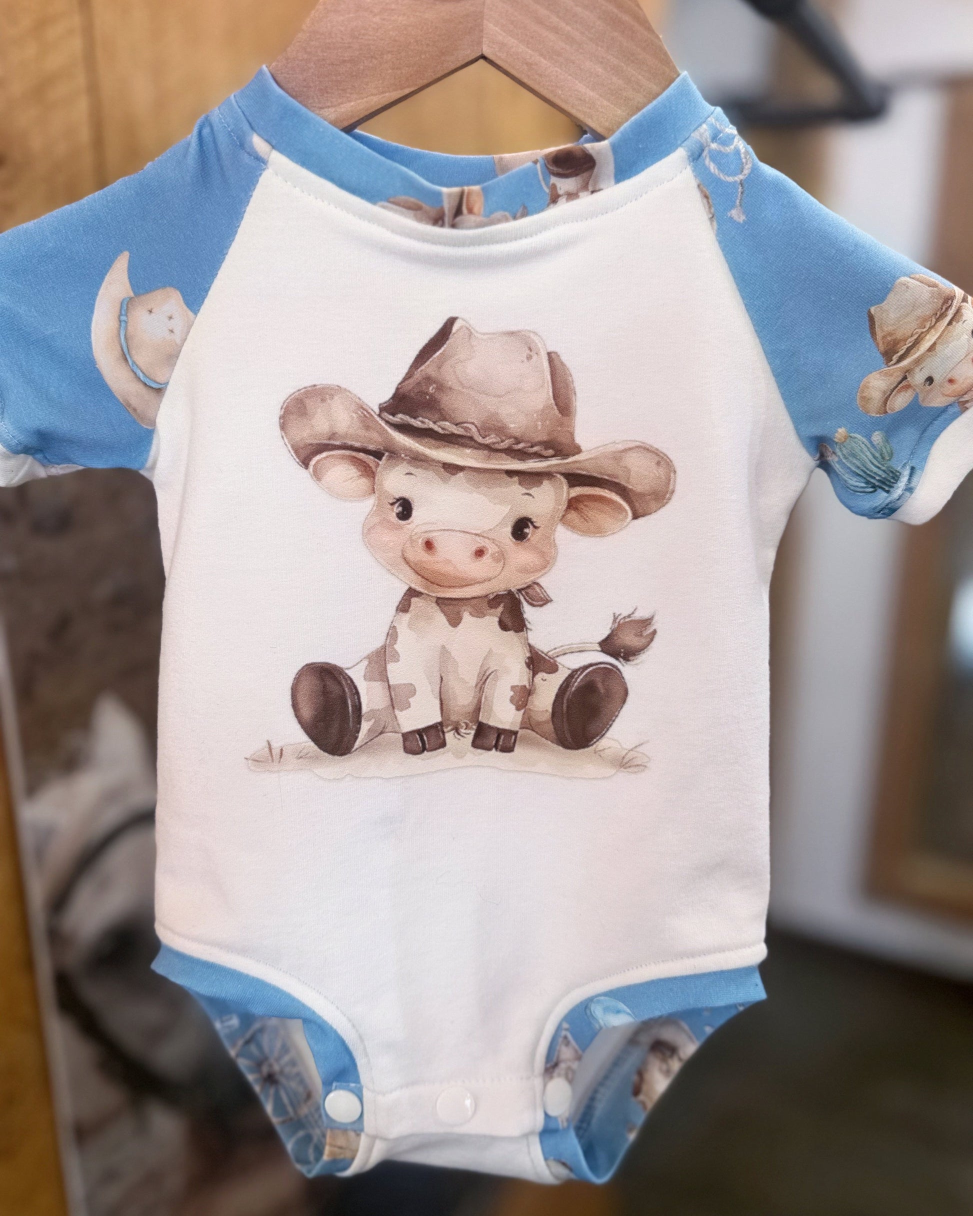 Baby onesie with cow design and blue sleeves on a hanger.