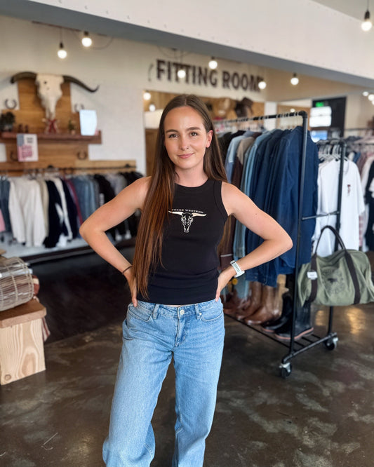 Woman in a black tank top and blue jeans standing in a clothing store with racks of clothes in the background.