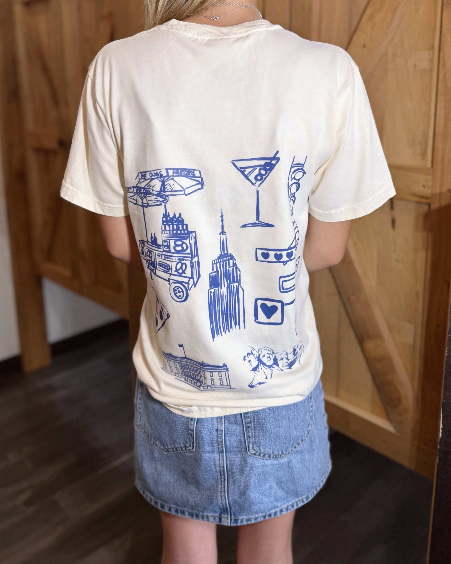 A young woman wears a cream-colored t-shirt with "Land of the Free Est. 1776" printed in black across the chest. The back showcases blue graphic illustrations of American icons, including the Empire State Building, pizza, and a food cart. She pairs it with a denim skirt and cowboy boots.