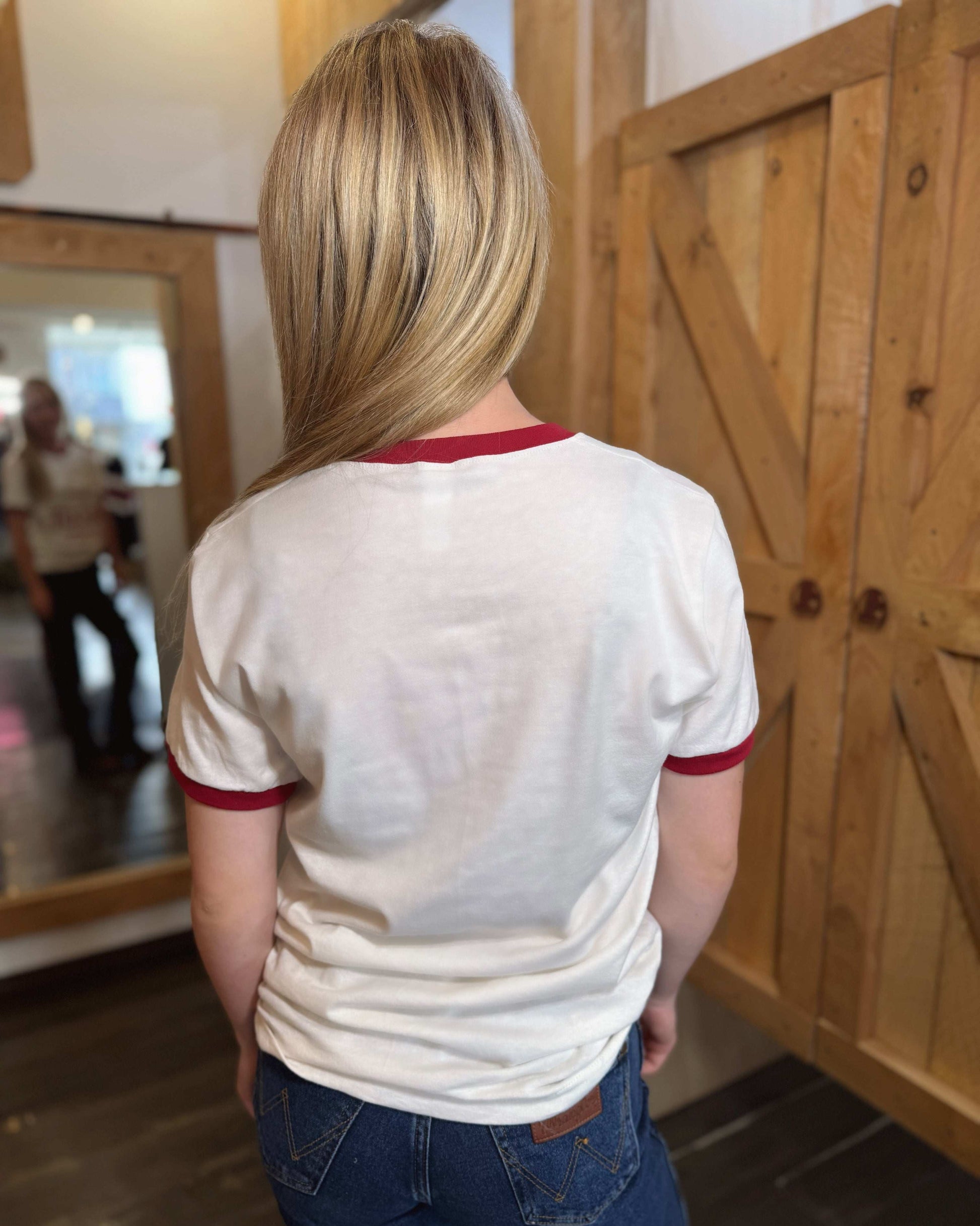 A young woman wears a white ringer t-shirt with red trim on the collar and sleeves. The front of the shirt features a red graphic with the words "Welcome to Tailgate City, Norman, Oklahoma," along with an outline of a cowboy hat. The t-shirt has a classic, slightly fitted cut. The back of the shirt is plain white with red trim. She pairs it with blue jeans and poses in front of a rustic wooden-paneled background.