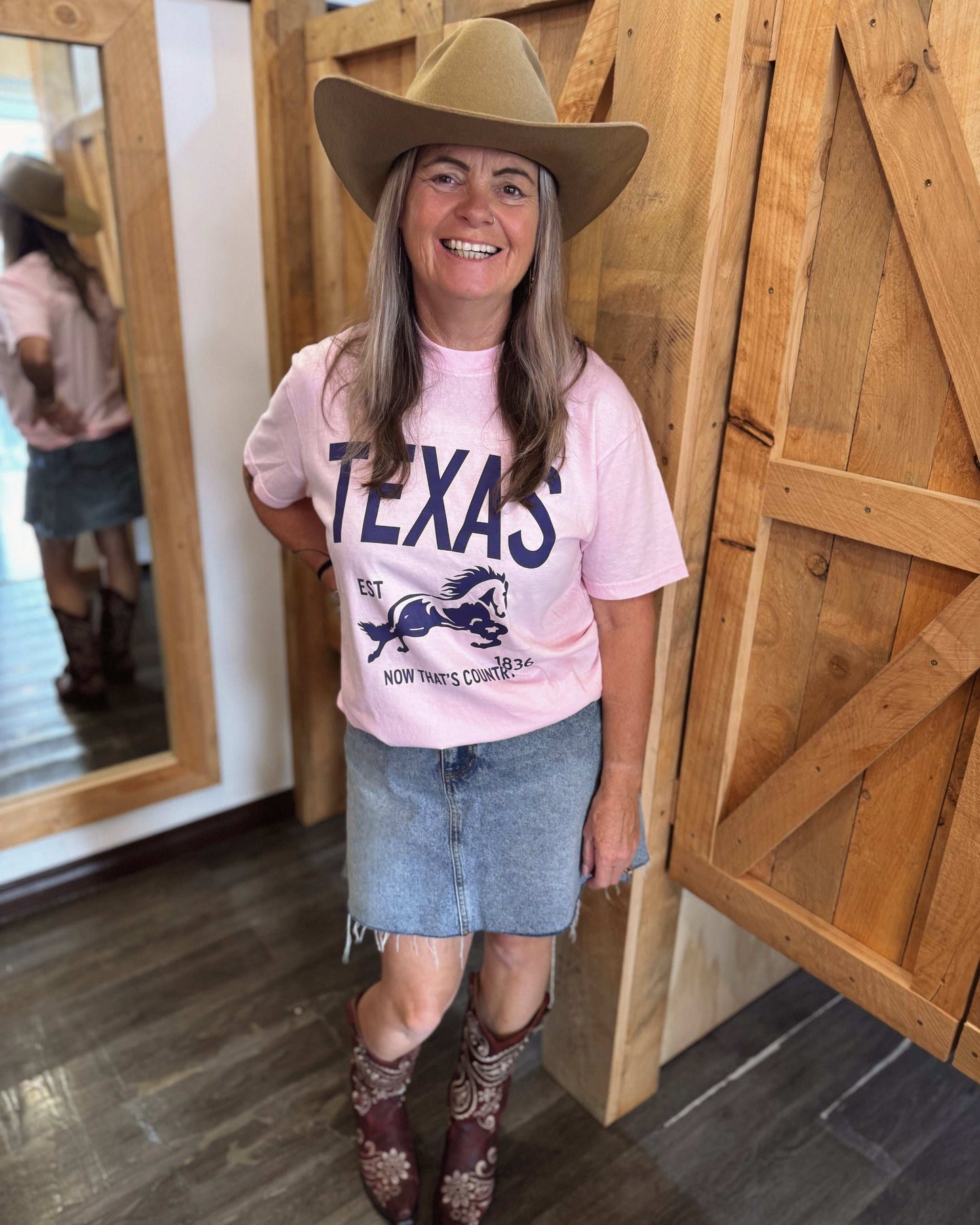 A woman with long, wavy hair wears a pastel pink oversized t-shirt with bold navy blue text that reads "TEXAS" along with a galloping horse graphic and the words "EST" and "Now That's Country." She pairs the shirt with a frayed denim mini skirt, a wide-brimmed beige cowboy hat, and embroidered cowboy boots. From the back, showing the plain pink back of the shirt with a relaxed fit.