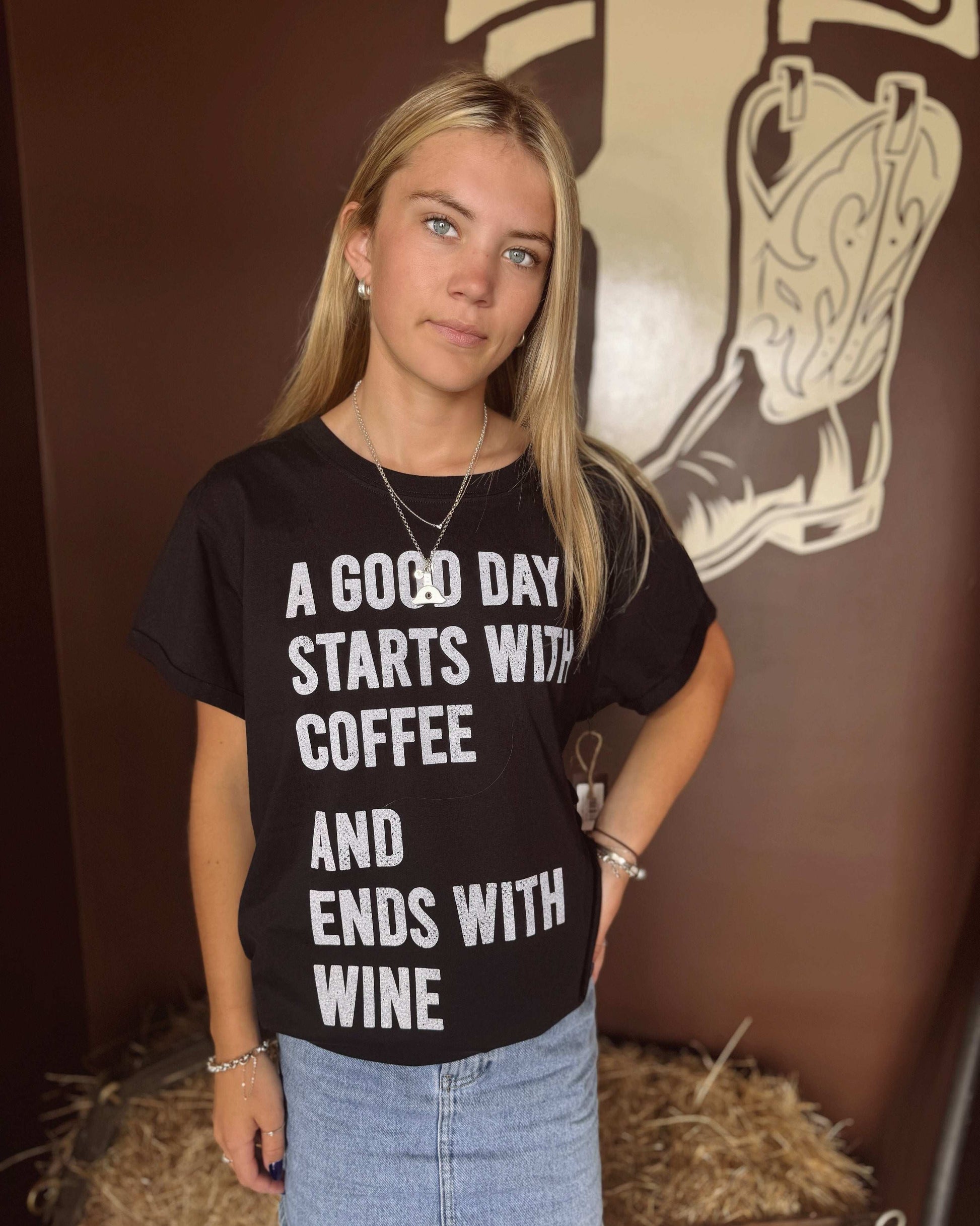  A woman with long blonde hair wears a black oversized graphic tee with bold white lettering that reads, "A GOOD DAY STARTS WITH COFFEE AND ENDS WITH WINE." The shirt features a relaxed fit with rolled-up sleeves and is paired with a light blue denim mini skirt. She is standing in front of a brown western-themed wall with a large boot illustration, giving the tee a rustic, country feel. In the second image, she is turned around to showcase the solid black back of the shirt, 