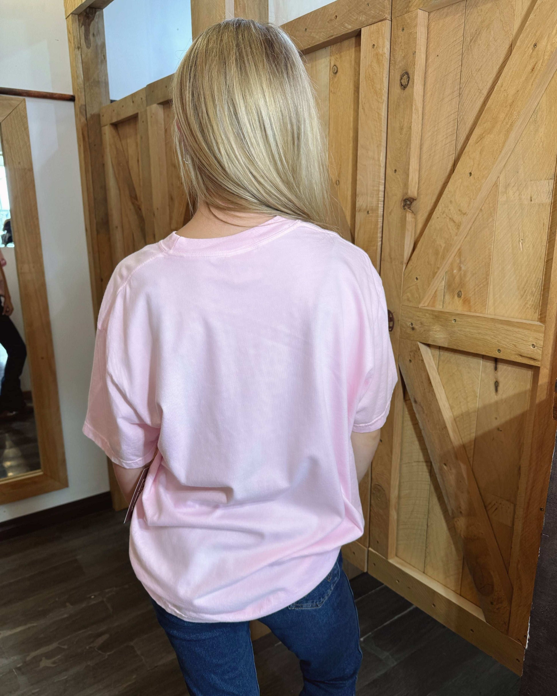 A pastel pink oversized T-shirt featuring a bold graphic print of a metallic gold cowboy hat with a pink and white ribbon tied around it. The shirt has a relaxed fit, short sleeves, and a classic crew neckline. The back of the shirt is plain pink. Modeled with dark blue jeans against a rustic wooden background.