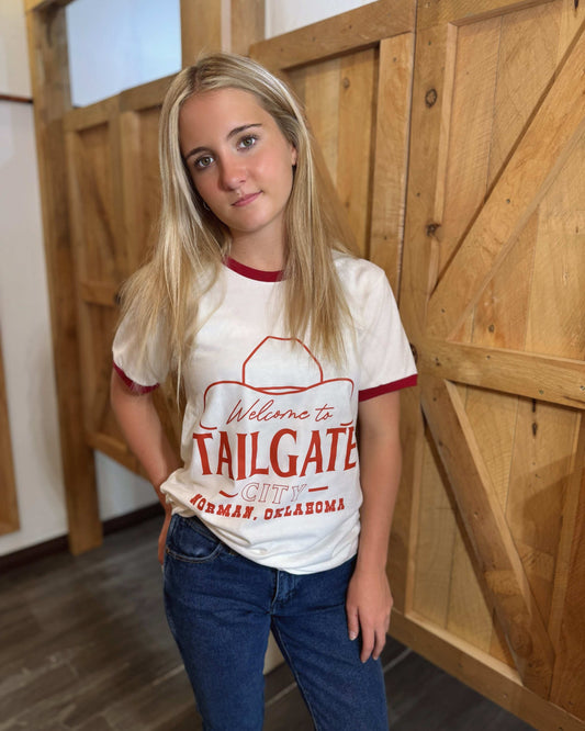 A young woman wears a white ringer t-shirt with red trim on the collar and sleeves. The front of the shirt features a red graphic with the words "Welcome to Tailgate City, Norman, Oklahoma," along with an outline of a cowboy hat. The t-shirt has a classic, slightly fitted cut. The back of the shirt is plain white with red trim. She pairs it with blue jeans and poses in front of a rustic wooden-paneled background.