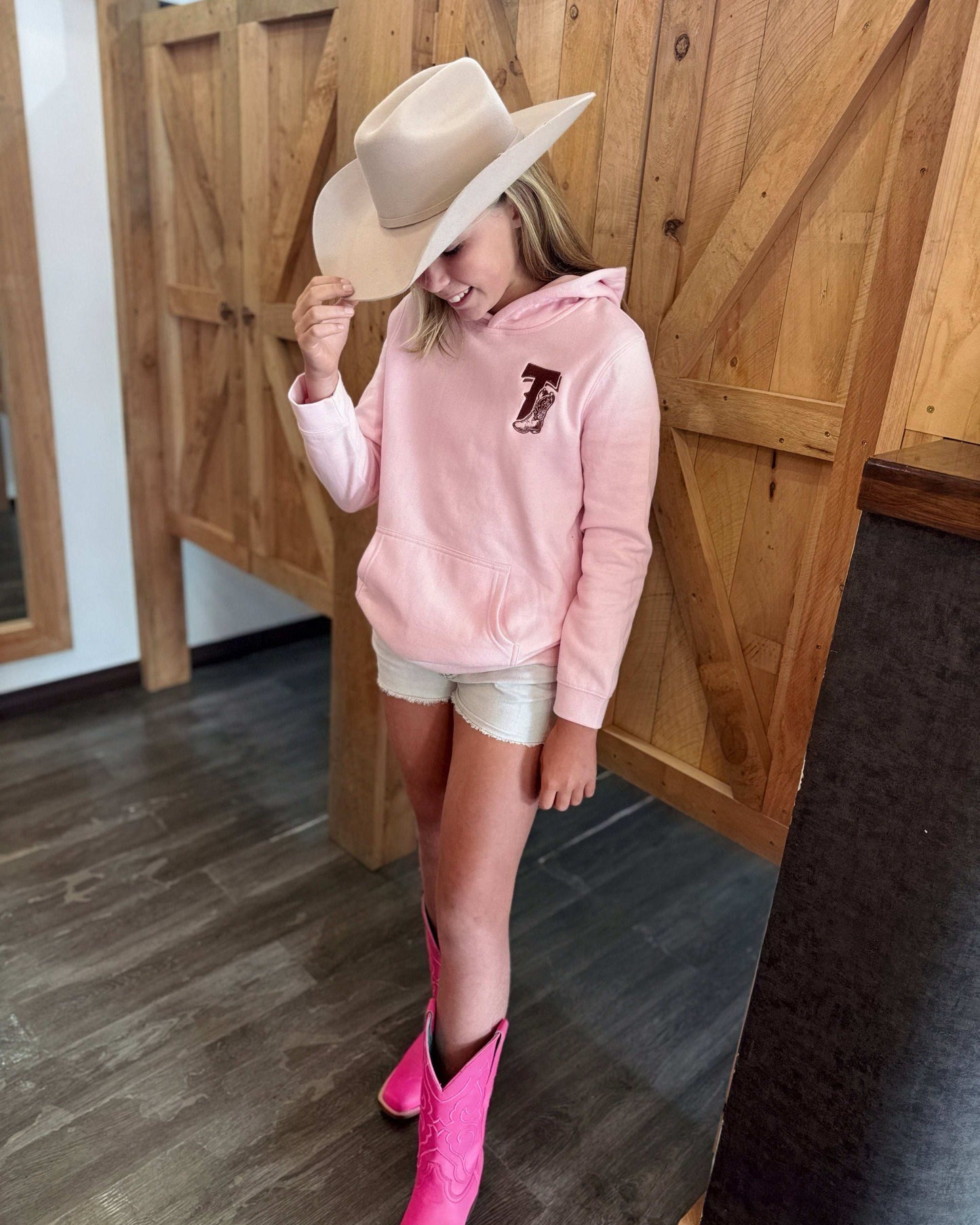 Pale pink Texas Western Apparel youth/kids hoodie with detailed embroidery. The front features a stitched 'T' and a Western-style boot, while the back displays the Texas Western Apparel logo. Perfect for little cowboys and cowgirls who love bold Western style!