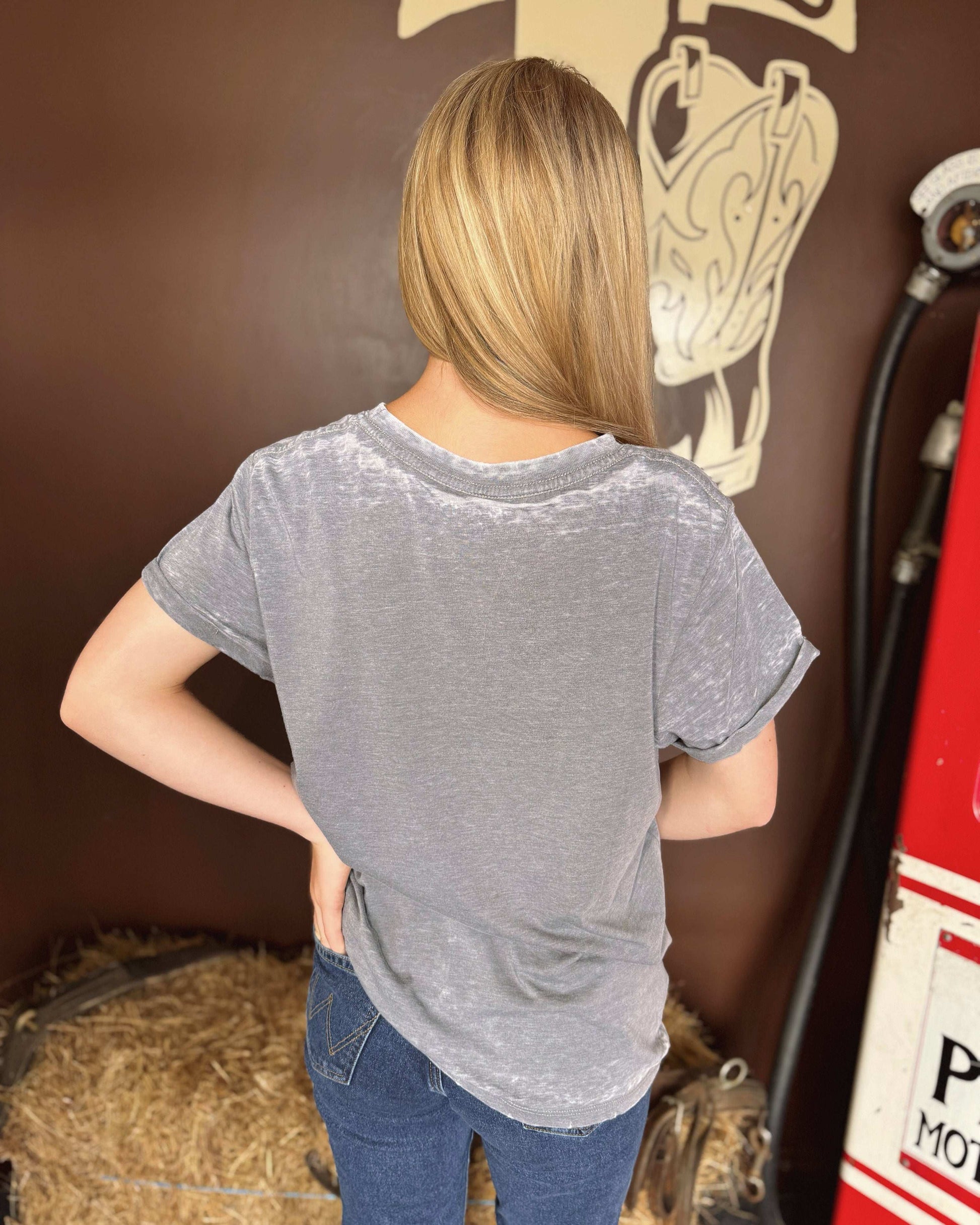 A young woman with straight blonde hair wears a faded gray t-shirt with a western-style graphic of a cowboy boot, a hat, a horseshoe, and the phrase "Saddle Up Cowgirl, Ride It Better" in orange and cream tones. She pairs the tee with classic blue jeans. The background features a brown wall with a large boot illustration, a vintage gas pump, and hay bales.