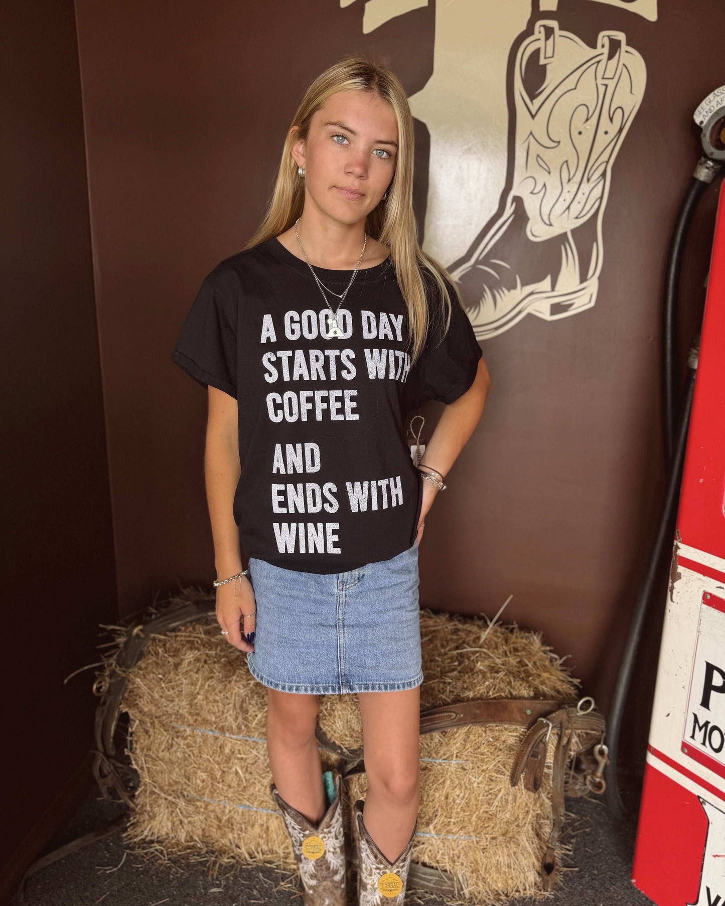  A woman with long blonde hair wears a black oversized graphic tee with bold white lettering that reads, "A GOOD DAY STARTS WITH COFFEE AND ENDS WITH WINE." The shirt features a relaxed fit with rolled-up sleeves and is paired with a light blue denim mini skirt. She is standing in front of a brown western-themed wall with a large boot illustration, giving the tee a rustic, country feel. In the second image, she is turned around to showcase the solid black back of the shirt, 