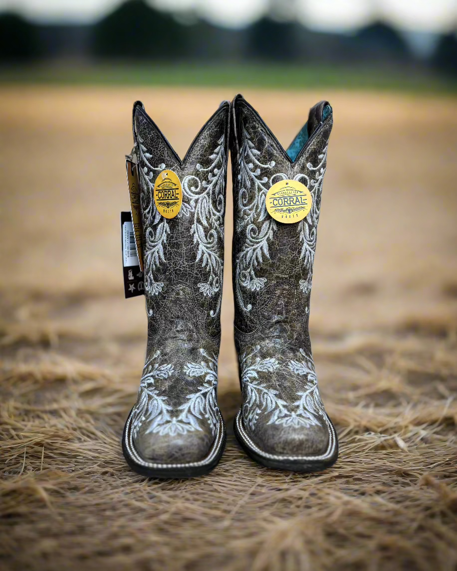 A pair of Corral cowboy boots with intricate white embroidery on distressed brown leather. The boots feature a classic western silhouette with a pointed toe, sturdy black heel, and a turquoise interior lining. The detailed floral embroidery not only adds an elegant touch but also features glow-in-the-dark white stitching, making these boots stand out both day and night. 