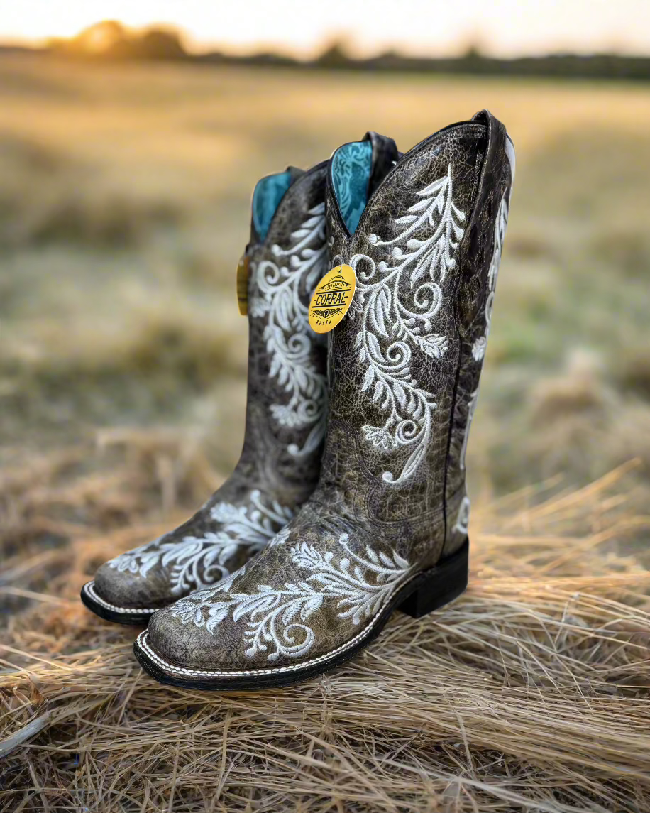 A pair of Corral cowboy boots with intricate white embroidery on distressed brown leather. The boots feature a classic western silhouette with a pointed toe, sturdy black heel, and a turquoise interior lining. The detailed floral embroidery not only adds an elegant touch but also features glow-in-the-dark white stitching, making these boots stand out both day and night. 