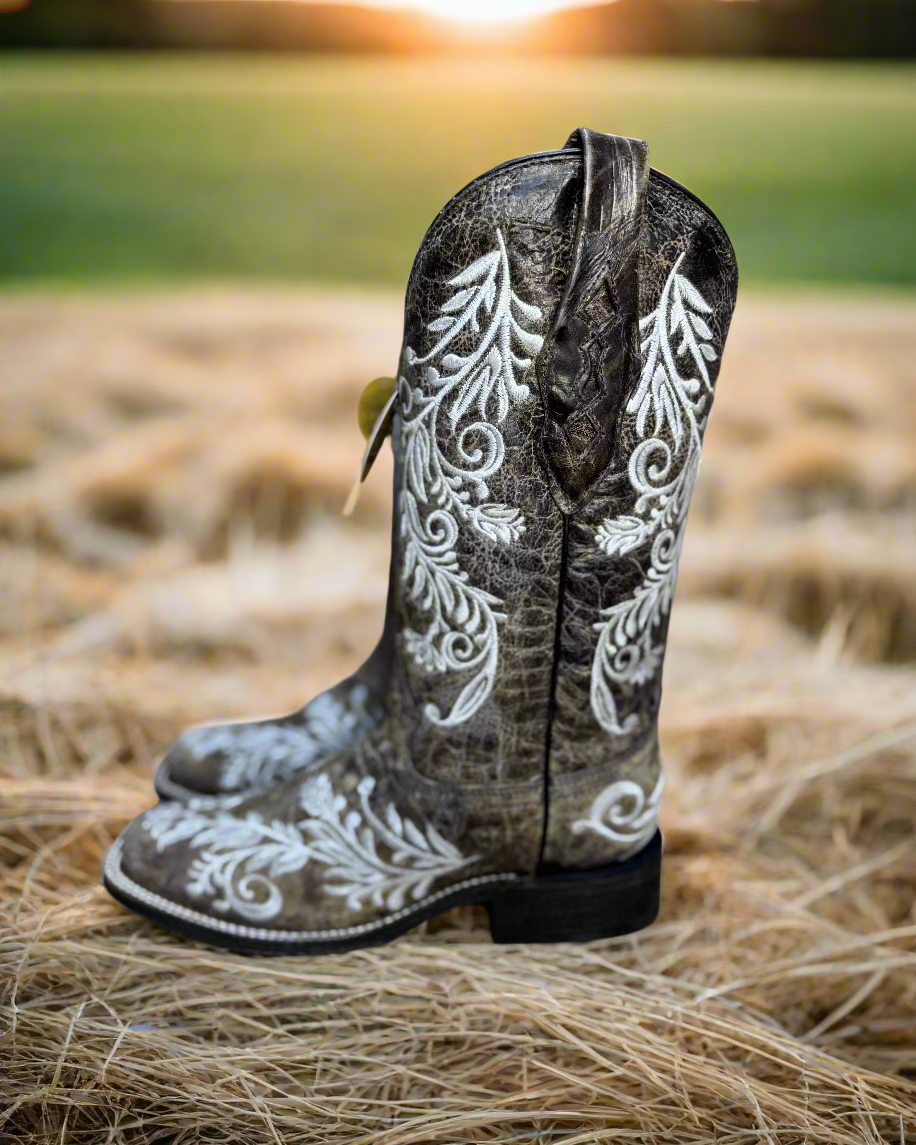 A pair of Corral cowboy boots with intricate white embroidery on distressed brown leather. The boots feature a classic western silhouette with a pointed toe, sturdy black heel, and a turquoise interior lining. The detailed floral embroidery not only adds an elegant touch but also features glow-in-the-dark white stitching, making these boots stand out both day and night. 