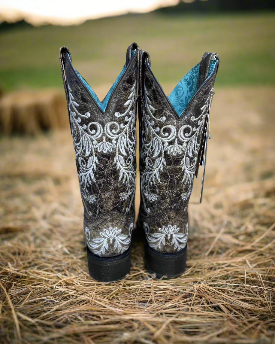 A pair of Corral cowboy boots with intricate white embroidery on distressed brown leather. The boots feature a classic western silhouette with a pointed toe, sturdy black heel, and a turquoise interior lining. The detailed floral embroidery not only adds an elegant touch but also features glow-in-the-dark white stitching, making these boots stand out both day and night. 