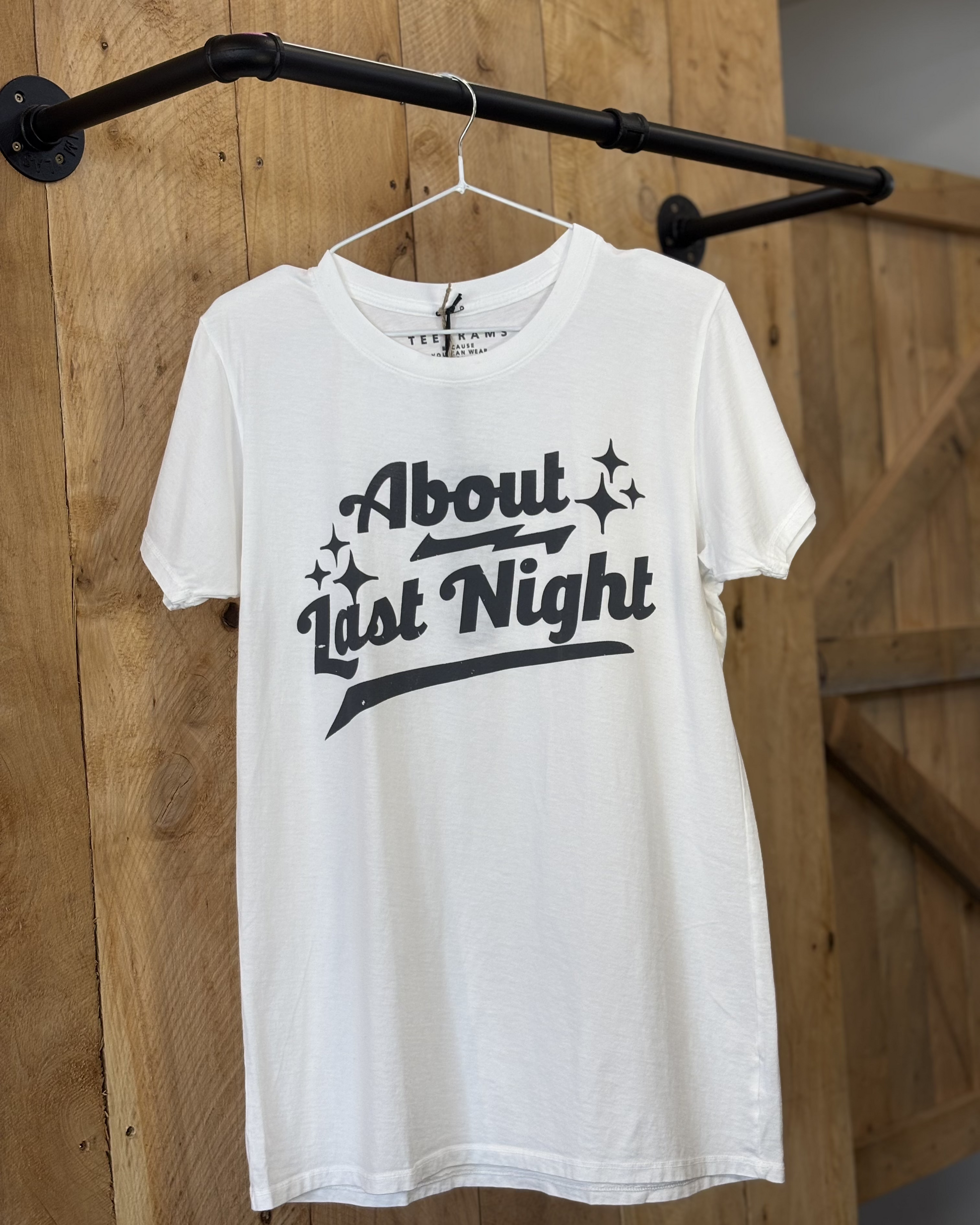 A young woman with long blonde hair wears a white graphic t-shirt with the phrase "About Last Night" in bold, black, retro-style lettering, accented with sparkles. She pairs the shirt with dark blue jeans and a black cowboy hat featuring a braided gold band. She is seated on a wooden bench in a rustic, western-themed setting with a "Hello Darling" pillow, a magazine, and black horseshoes mounted on the wooden wall behind her.