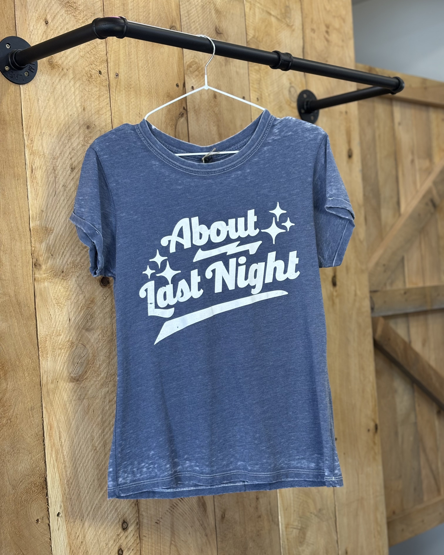 A young woman wears a heathered blue graphic tee with bold white lettering that reads "About Last Night," accented by decorative stars. She pairs the tee with a denim mini skirt and a black cowboy hat, embracing a western-inspired look. The tee’s casual fit and soft fabric make it a stylish yet comfortable choice for any country-loving wardrobe.