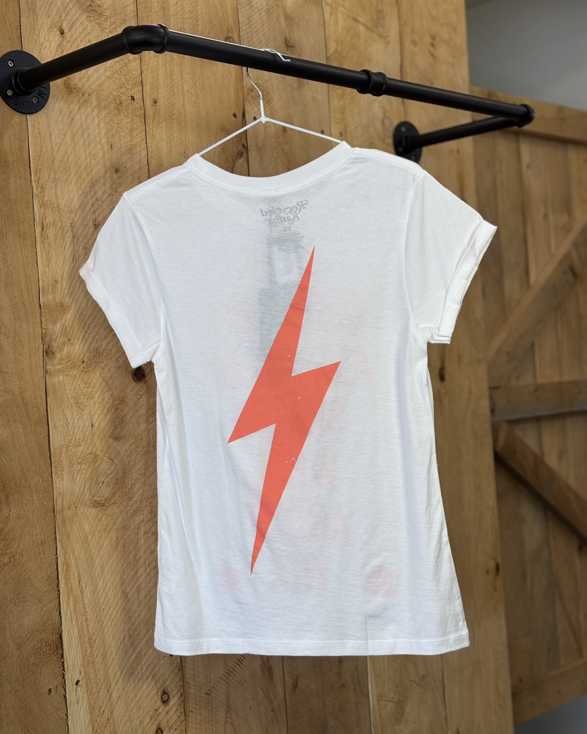 A woman wears a white graphic tee featuring bold red-orange text that reads, "AND SUDDENLY, ALL THESE SONGS WERE ABOUT YOU," with two stars at the bottom. The shirt has a relaxed fit and short sleeves, paired with classic blue jeans. The back of the tee showcases a striking red-orange lightning bolt design. The setting includes a rustic western-inspired backdrop with hay bales and vintage decor, complementing the casual country style.