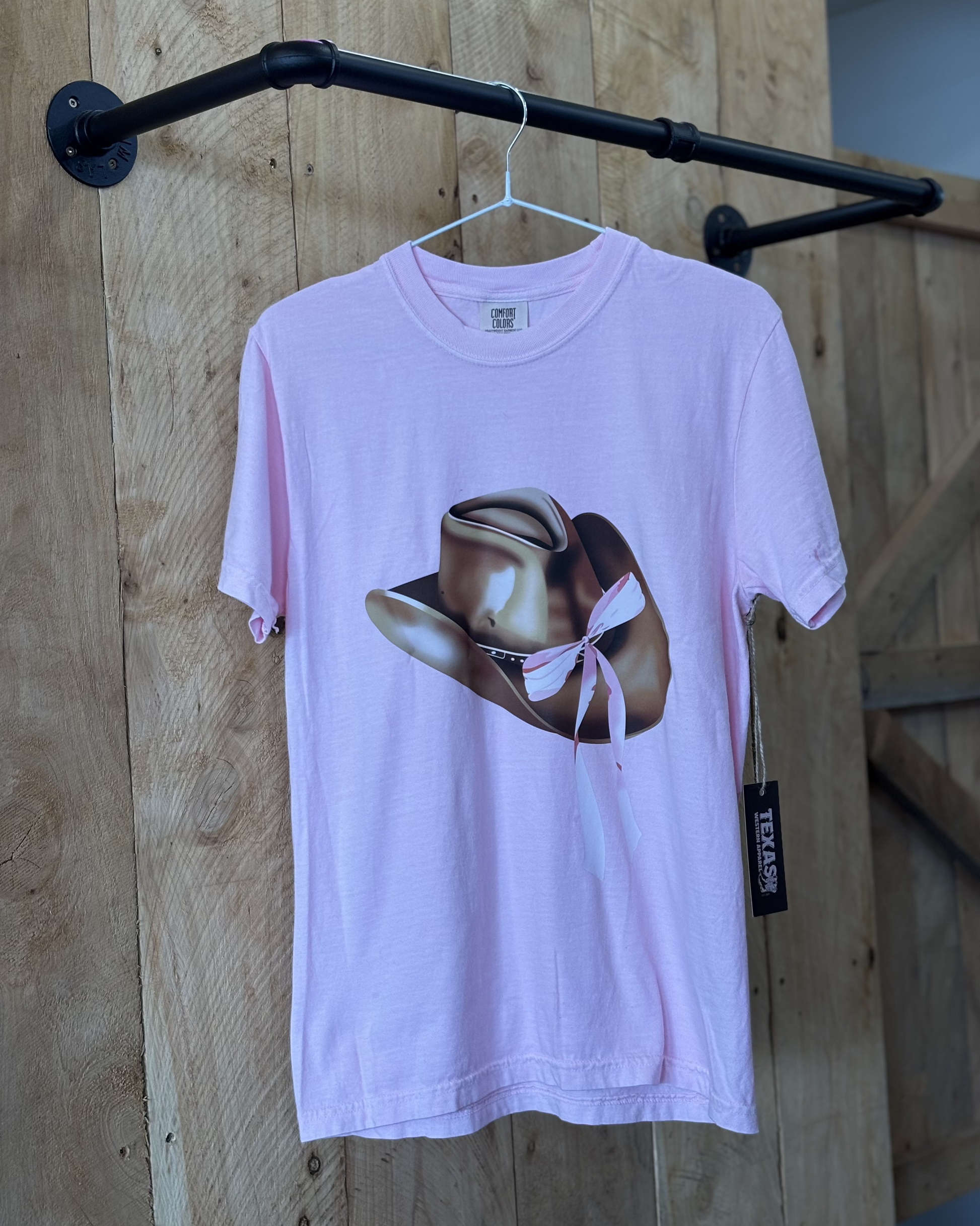 A pastel pink oversized T-shirt featuring a bold graphic print of a metallic gold cowboy hat with a pink and white ribbon tied around it. The shirt has a relaxed fit, short sleeves, and a classic crew neckline. The back of the shirt is plain pink. Modeled with dark blue jeans against a rustic wooden background.