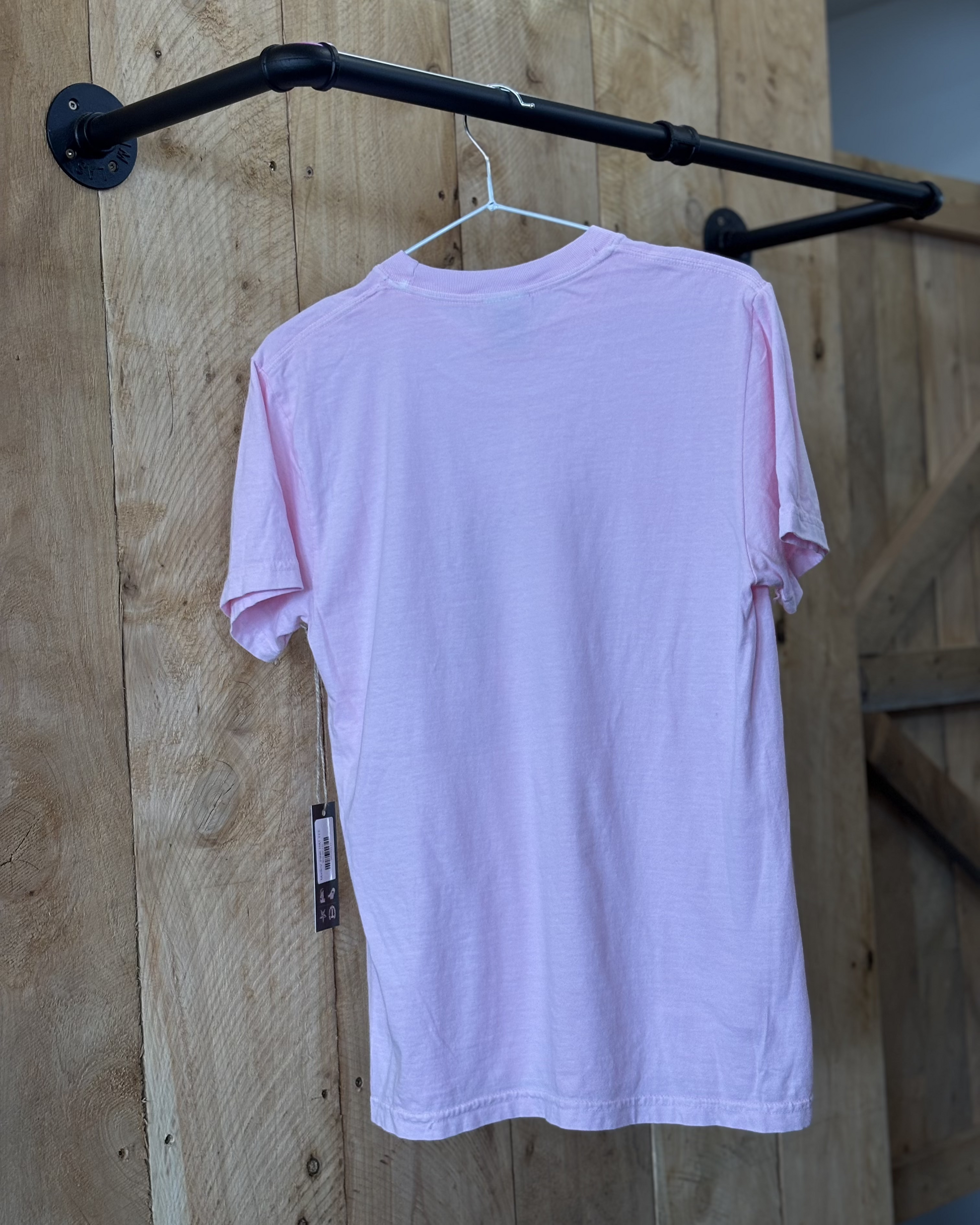 A pastel pink oversized T-shirt featuring a bold graphic print of a metallic gold cowboy hat with a pink and white ribbon tied around it. The shirt has a relaxed fit, short sleeves, and a classic crew neckline. The back of the shirt is plain pink. Modeled with dark blue jeans against a rustic wooden background.