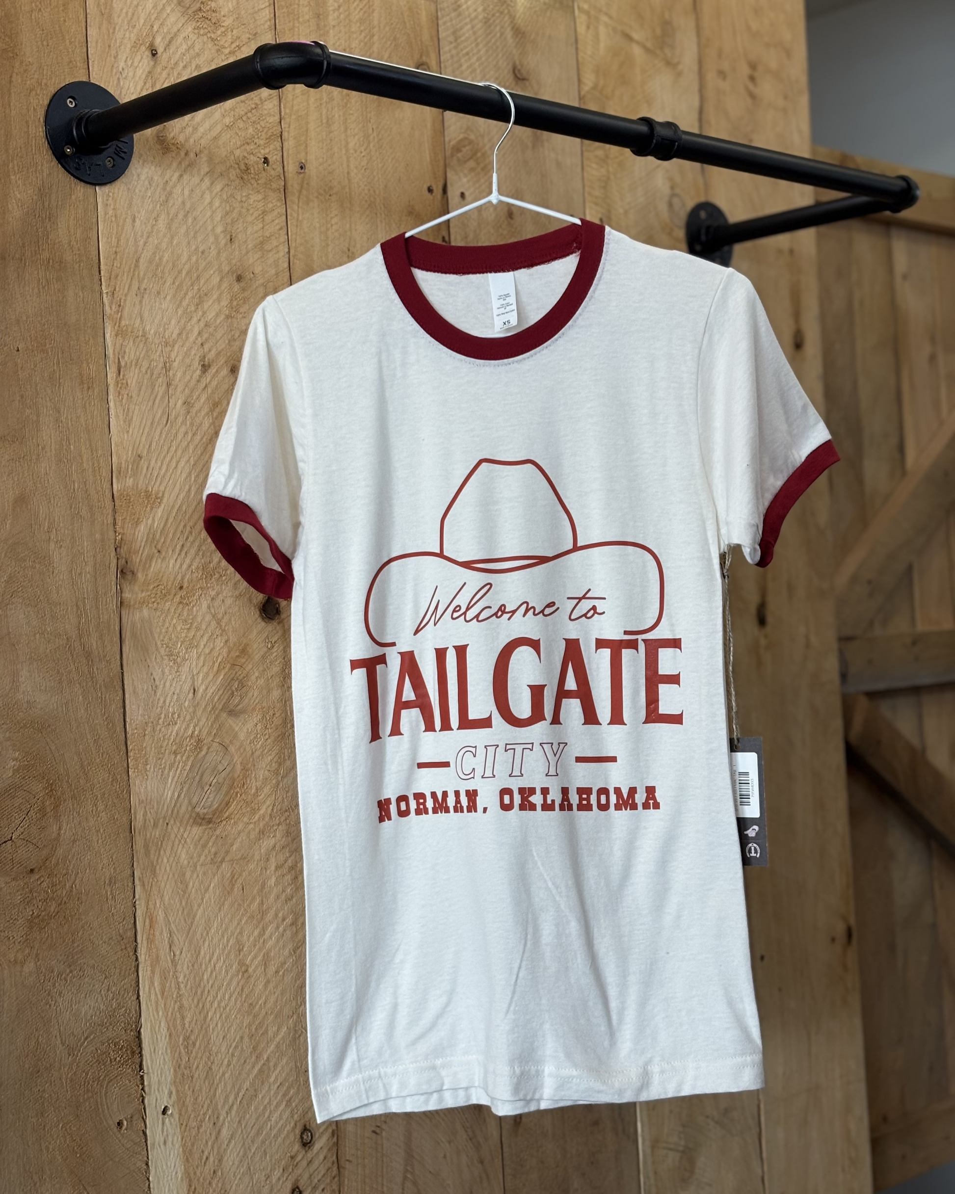 A young woman wears a white ringer t-shirt with red trim on the collar and sleeves. The front of the shirt features a red graphic with the words "Welcome to Tailgate City, Norman, Oklahoma," along with an outline of a cowboy hat. The t-shirt has a classic, slightly fitted cut. The back of the shirt is plain white with red trim. She pairs it with blue jeans and poses in front of a rustic wooden-paneled background.