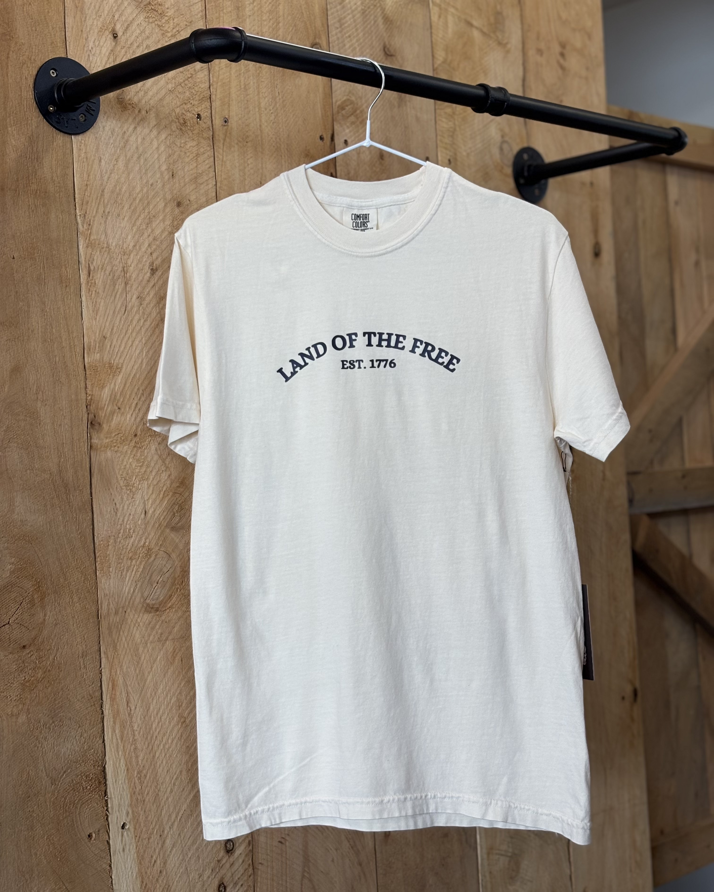 A young woman wears a cream-colored t-shirt with "Land of the Free Est. 1776" printed in black across the chest. The back showcases blue graphic illustrations of American icons, including the Empire State Building, pizza, and a food cart. She pairs it with a denim skirt and cowboy boots.