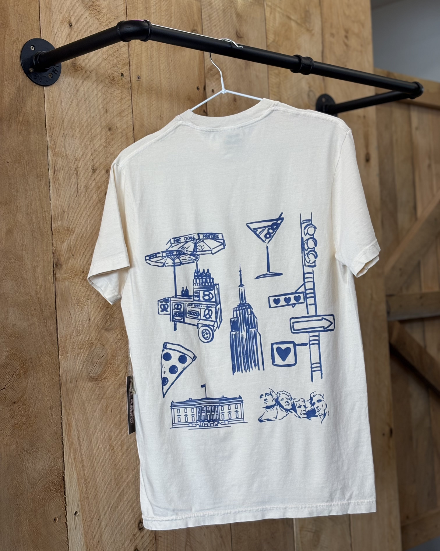 A young woman wears a cream-colored t-shirt with "Land of the Free Est. 1776" printed in black across the chest. The back showcases blue graphic illustrations of American icons, including the Empire State Building, pizza, and a food cart. She pairs it with a denim skirt and cowboy boots.