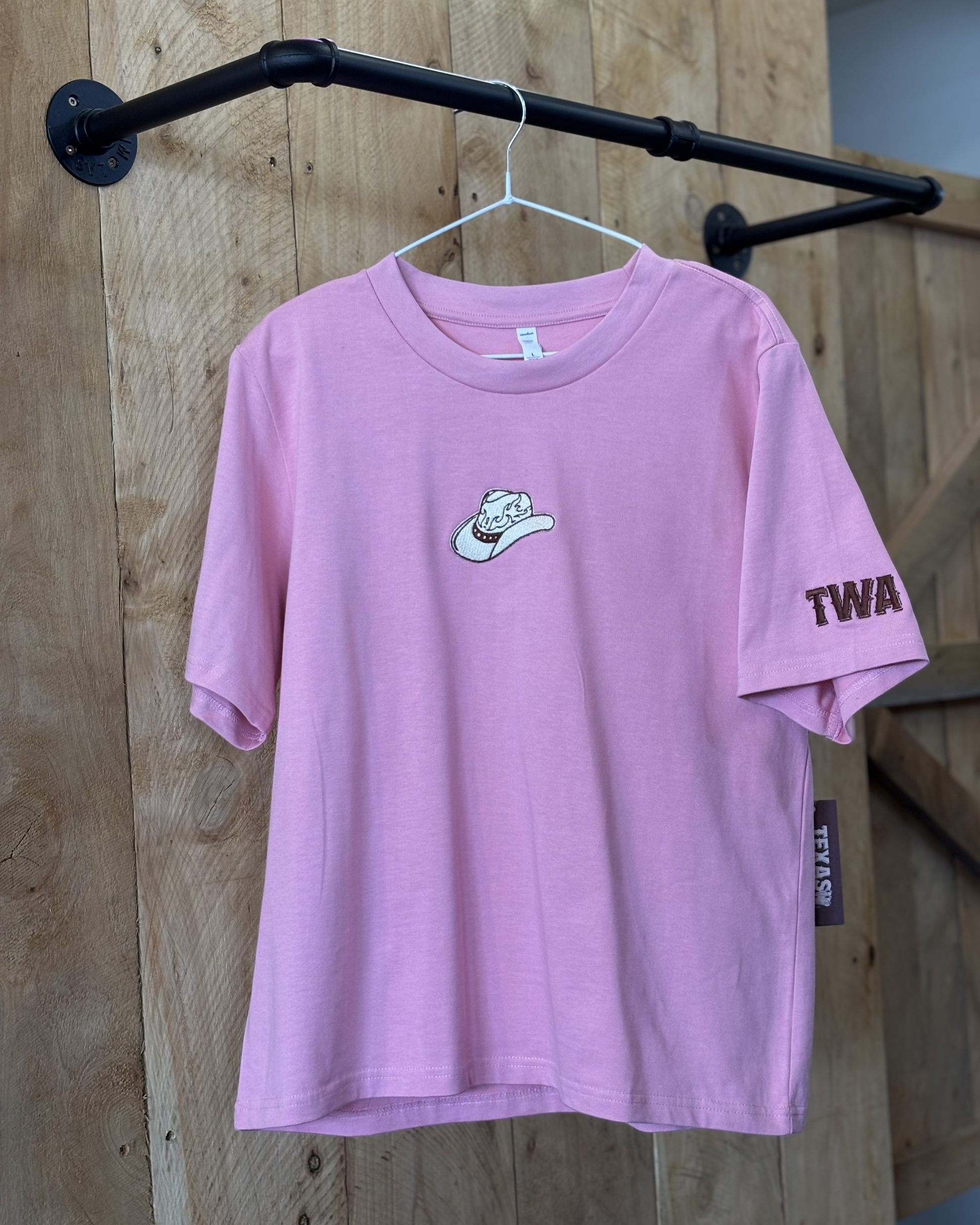 A pink women's Texas Western Apparel T-shirt with a classic western style. The front features an embroidered cowboy hat, while the sleeve is detailed with "TWA" initials, adding a subtle branded touch.