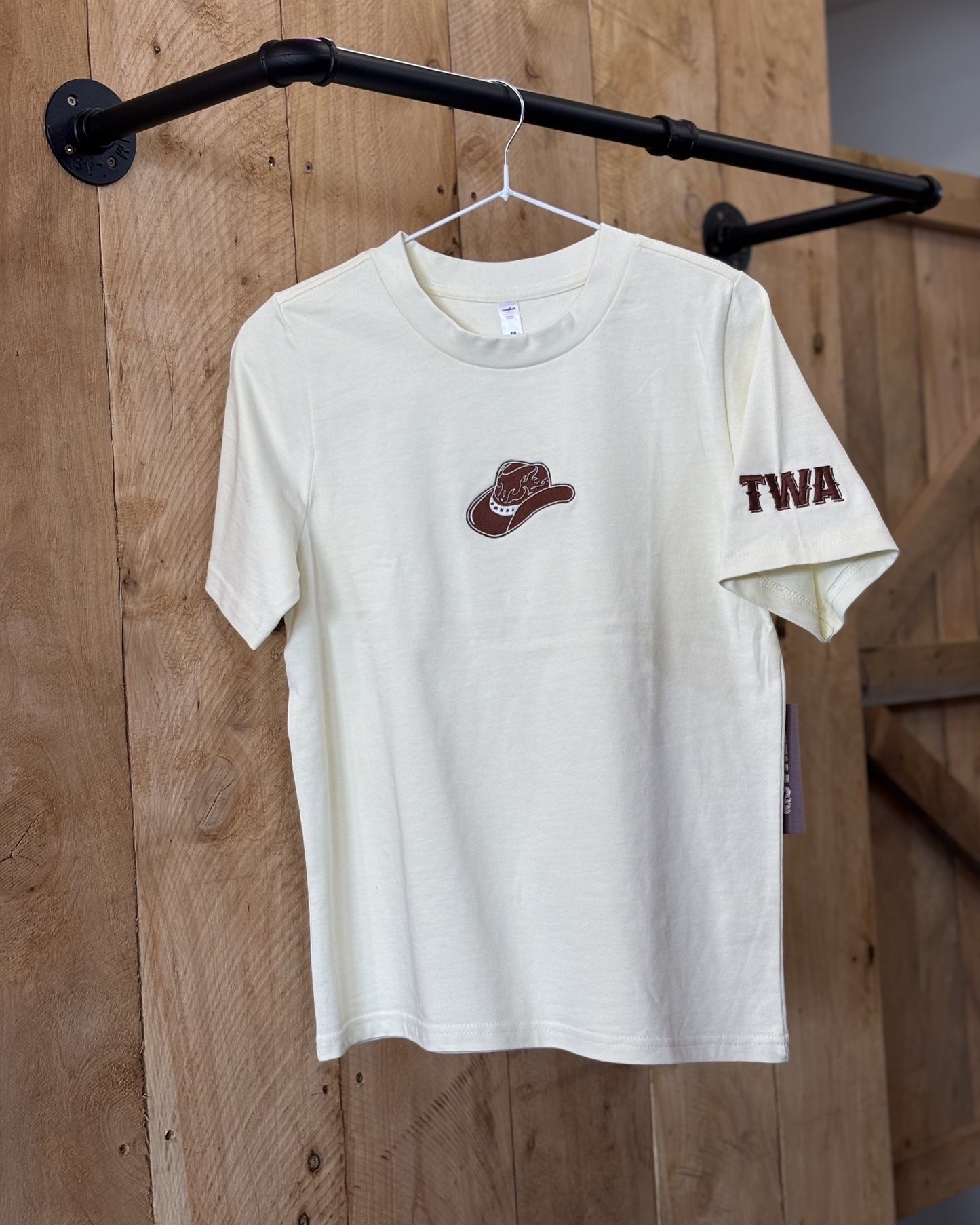 A butter yellow women's Texas Western Apparel T-shirt with a classic western style. The front features an embroidered cowboy hat, while the sleeve is detailed with "TWA" initials, adding a subtle branded touch.