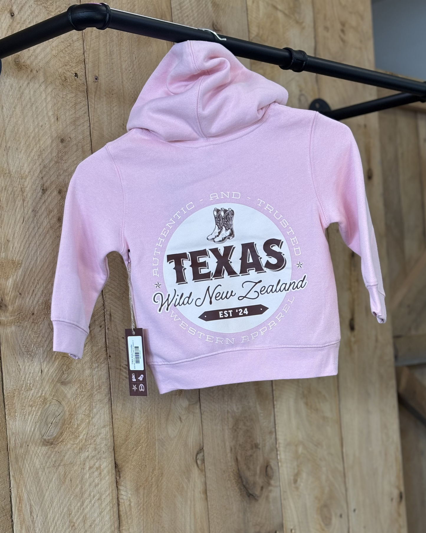 Pale pink Texas Western Apparel youth/kids hoodie with detailed embroidery. The front features a stitched 'T' and a Western-style boot, while the back displays the Texas Western Apparel logo. Perfect for little cowboys and cowgirls who love bold Western style!