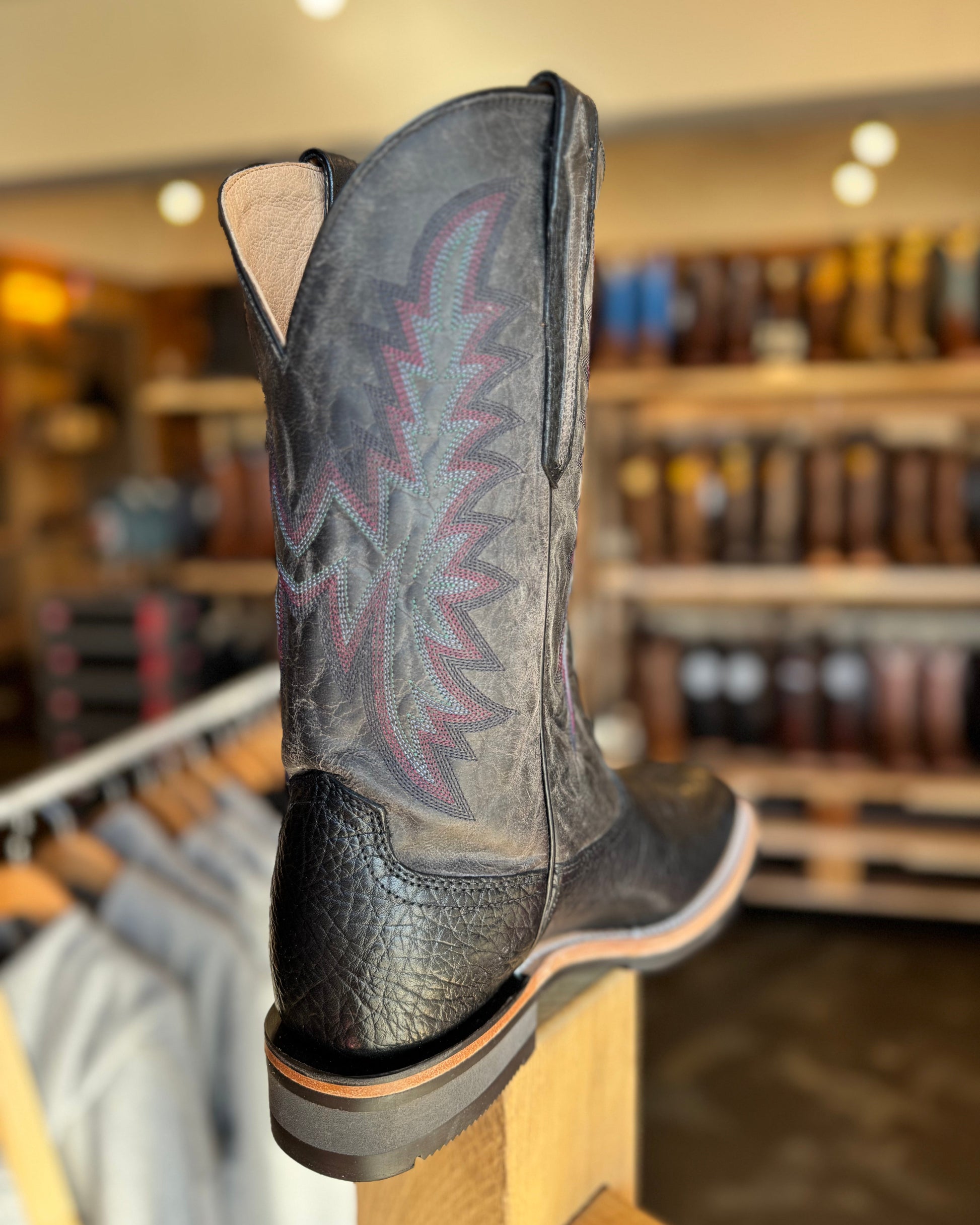 Black cowboy boot with intricate design on a wooden display stand in a store setting.
