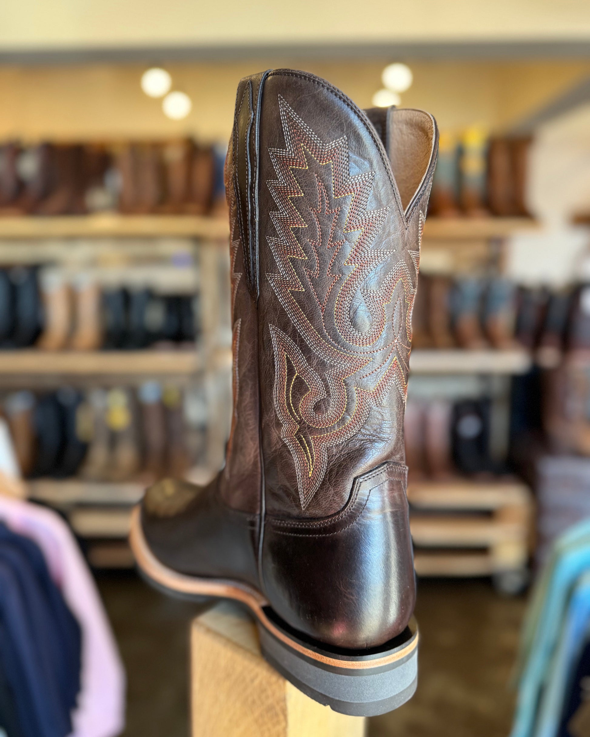 Brown cowboy boot with intricate stitching on a wooden block in a store setting.