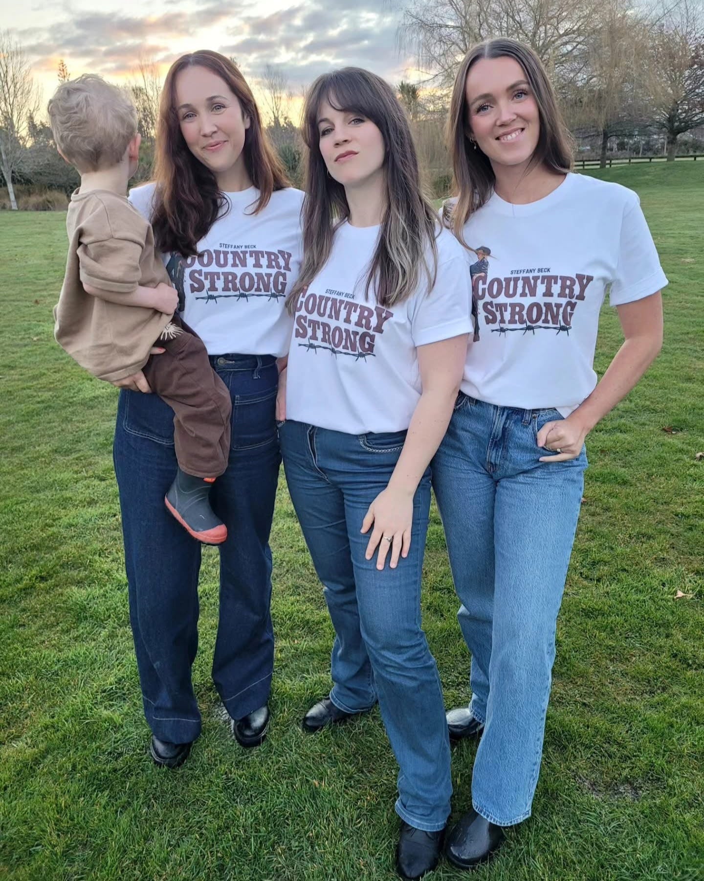Four people wearing 'Country Strong' t-shirts standing on grass with a sunset in the background.