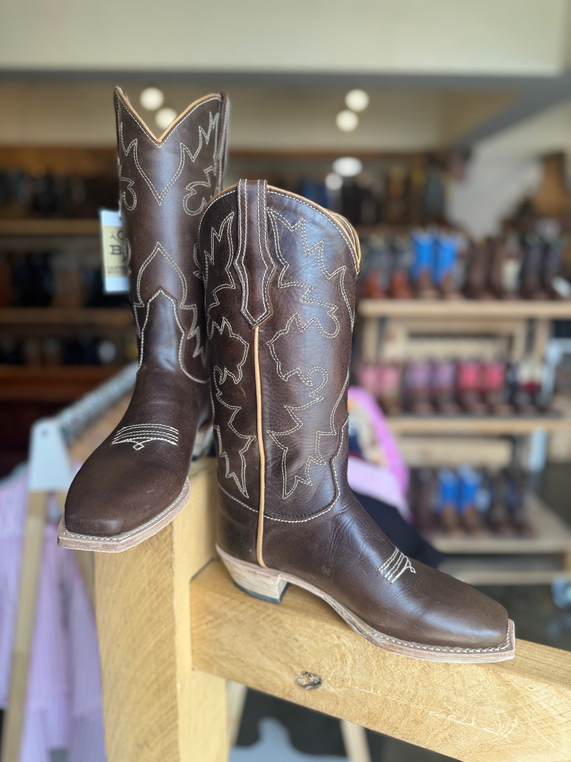 Pair of brown cowboy boots with intricate designs on a wooden block in a store setting.